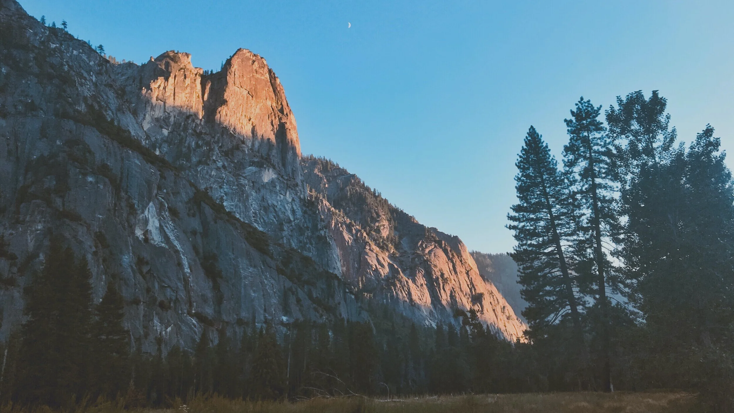 Sunlit rugged mountain cliffs with tall pine trees in foreground, under a clear blue sky with a crescent moon.