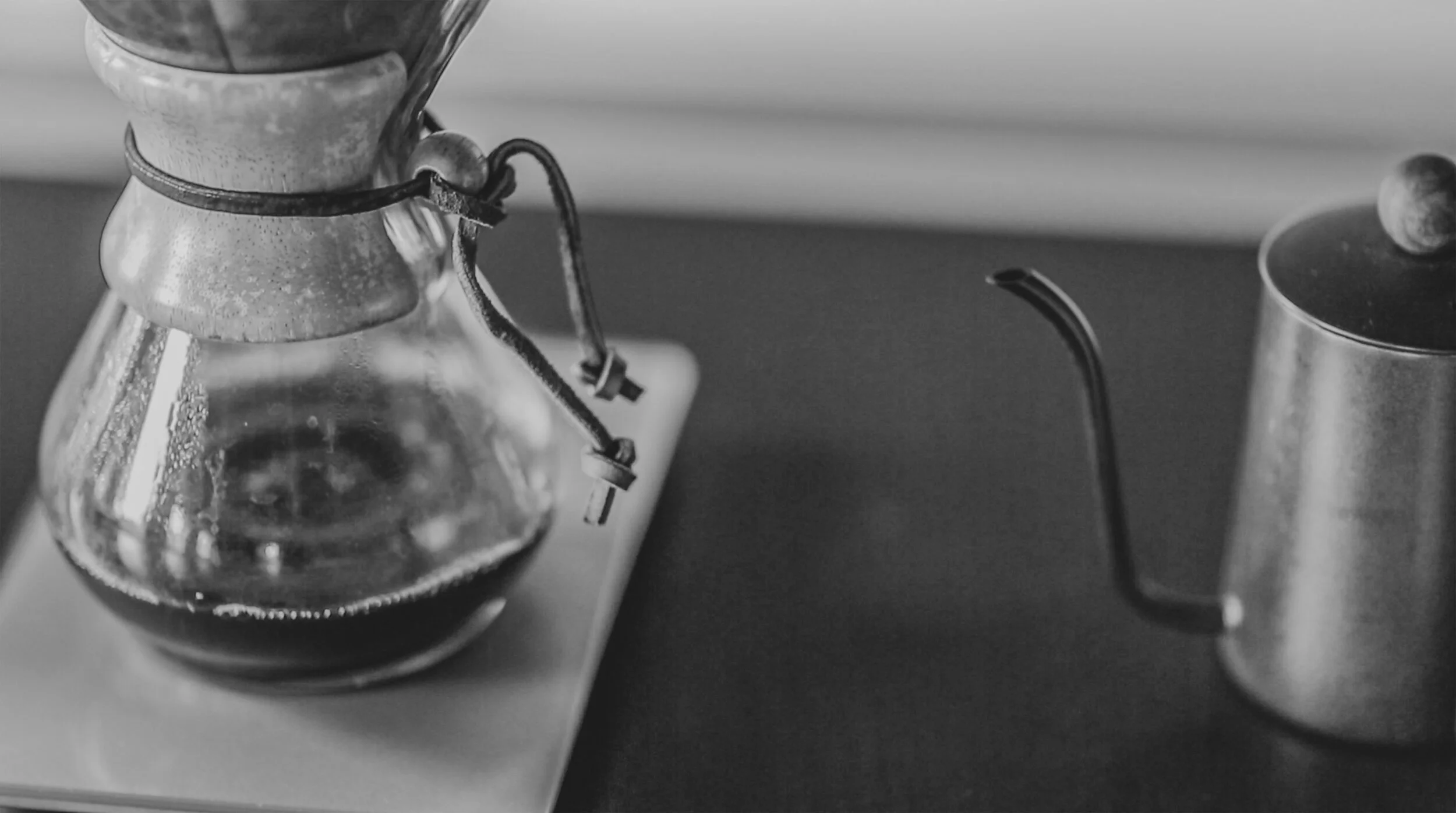 Black and white photo of a glass coffee pot on a scale, next to a metal kettle with a spout and lid, on a dark surface.