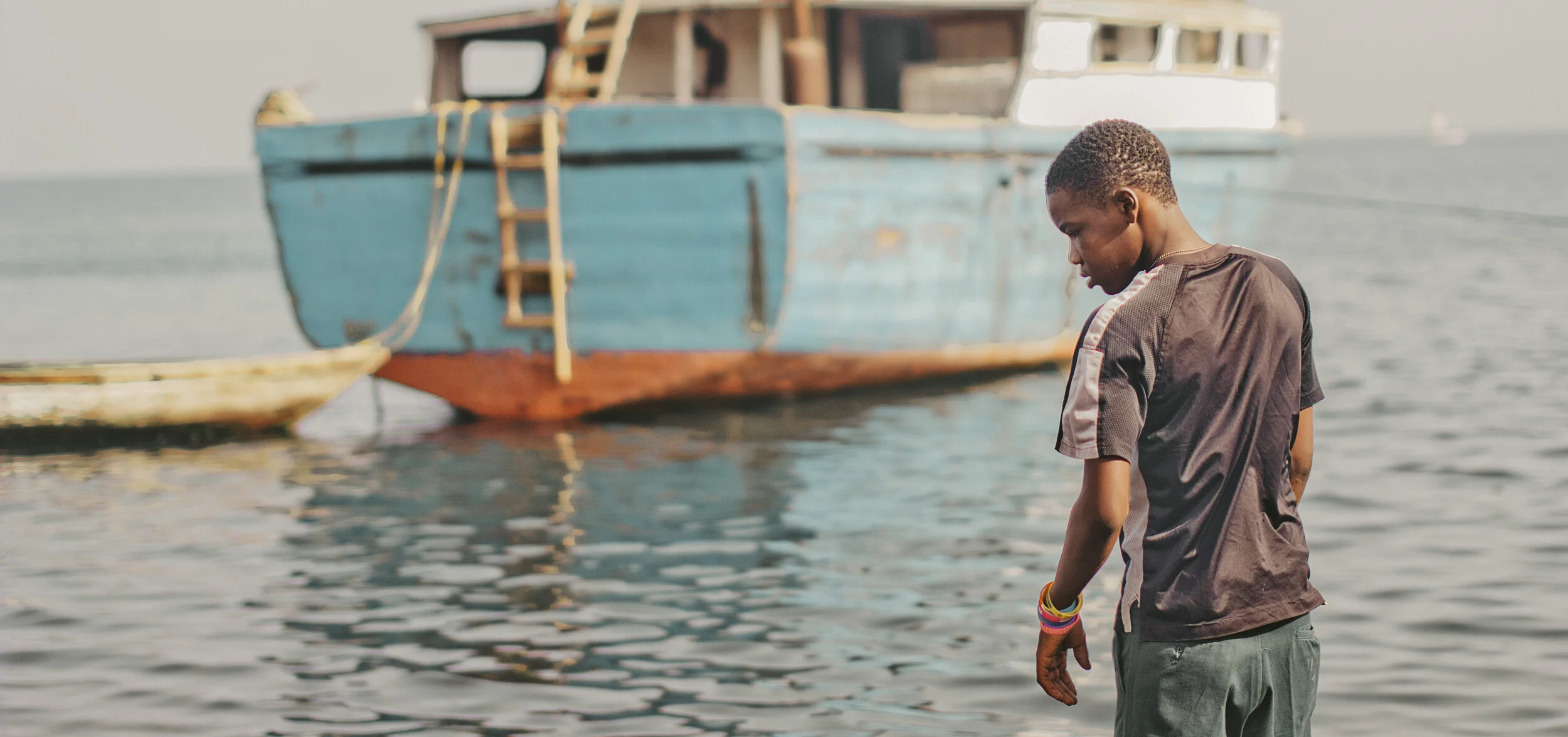 A young boy standing by the water's edge near a large boat, looking down with a contemplative expression.