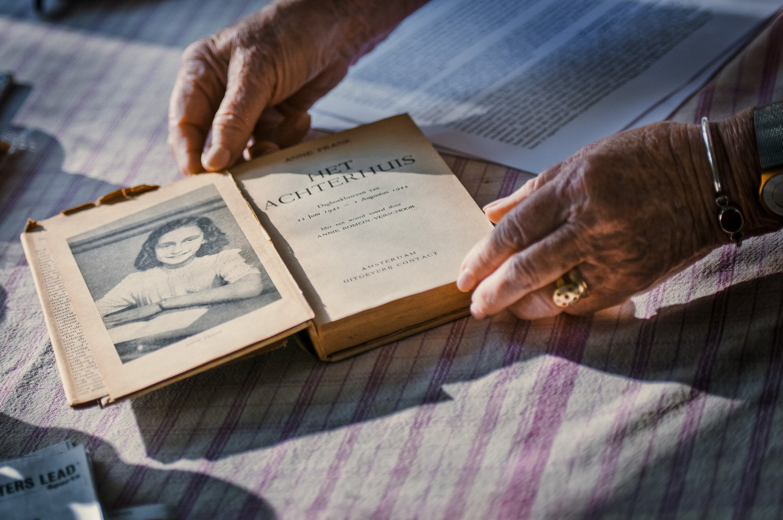 A person with elderly hands is flipping through an old book with a black and white portrait of a young girl on the left page and text on the right page, on a table with a checkered tablecloth.