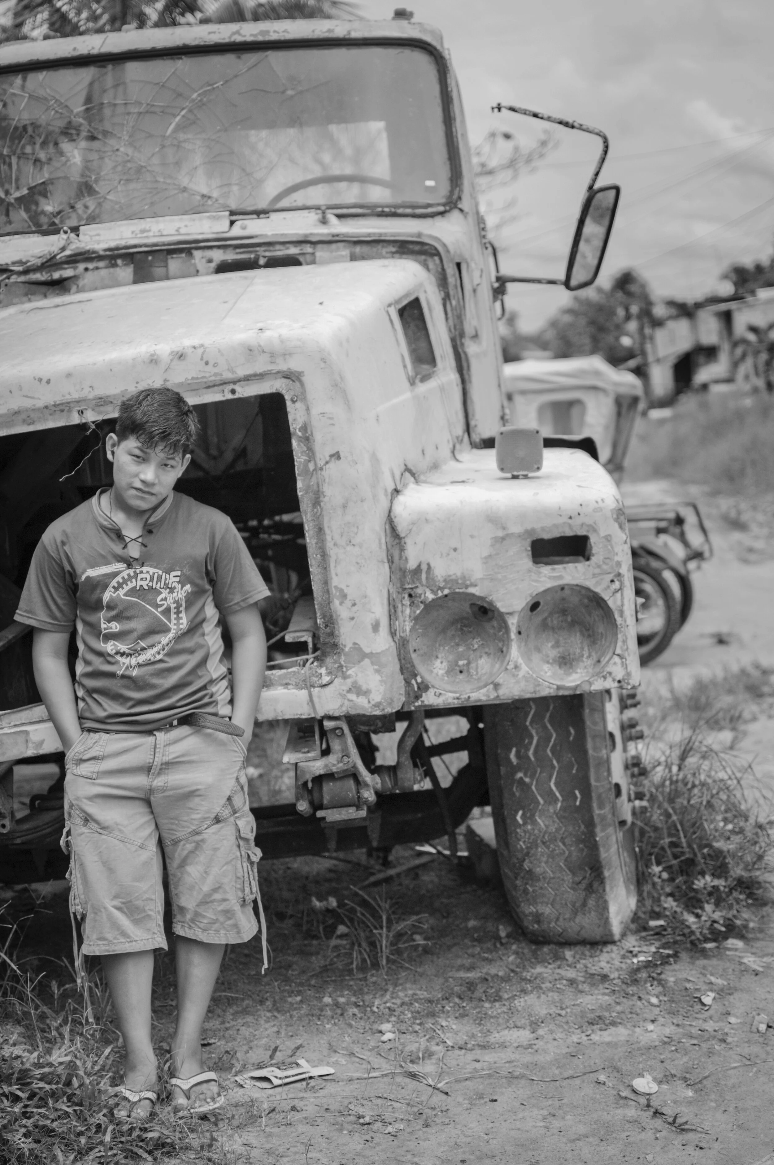 A boy standing next to an old, weathered truck in a rural area, with more trucks in the background.