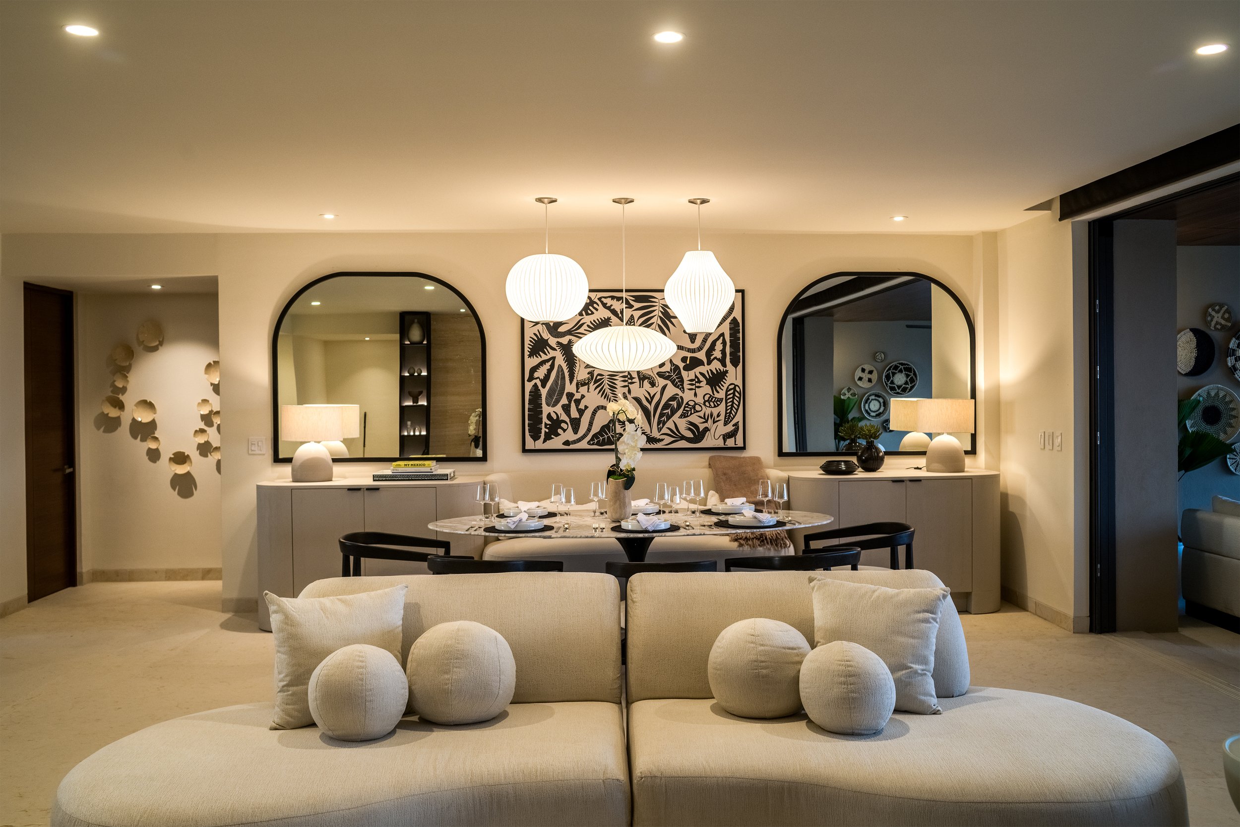 A residential space designed in warm neutrals.  The image is of the living room sofa and also encompasses the dining room table and banquette.  There are beautiful light fixtures as well as many accessories.