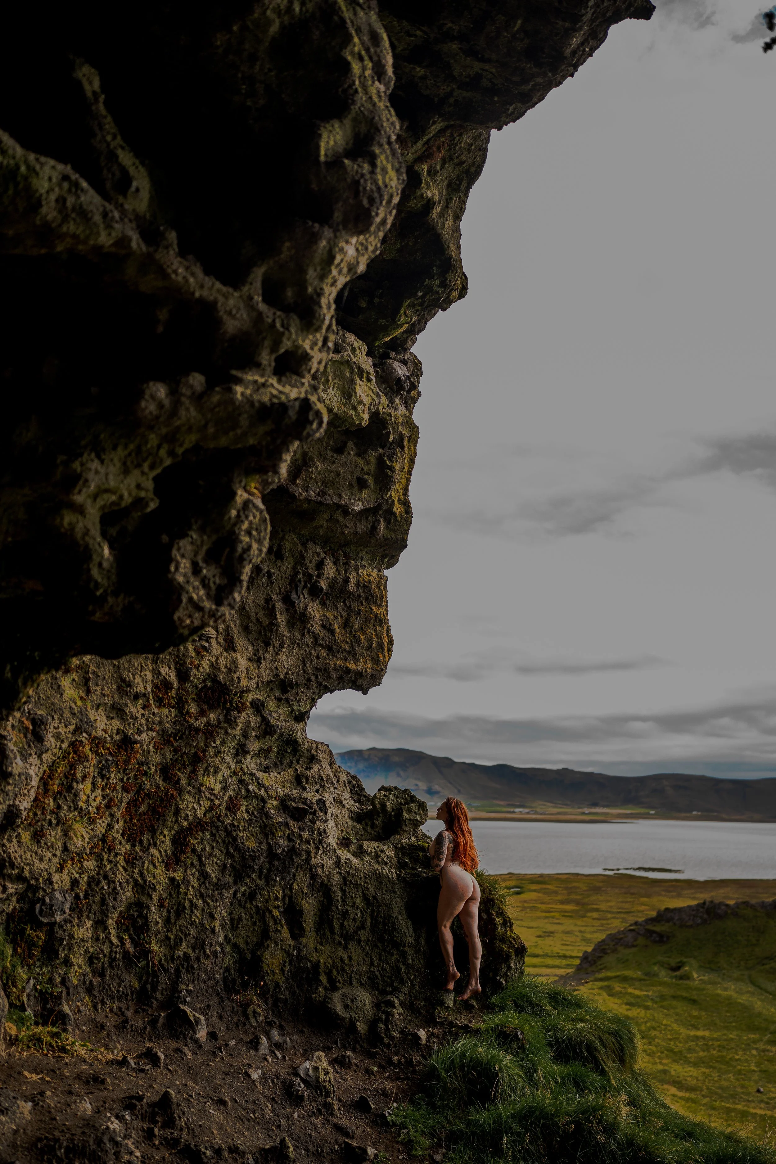 woman  with red hair standing on a rocky cliff overlooking a scenic landscape with green fields, lake, and mountains in the distance in iceland during a boudoir photography session