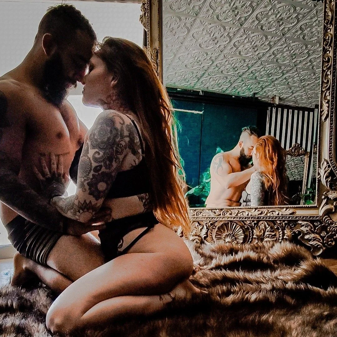A couple kneeling on a fur rug, embracing and touching foreheads, reflected in an ornate mirror. The woman has long hair and tattoos, and both are partially dressed.