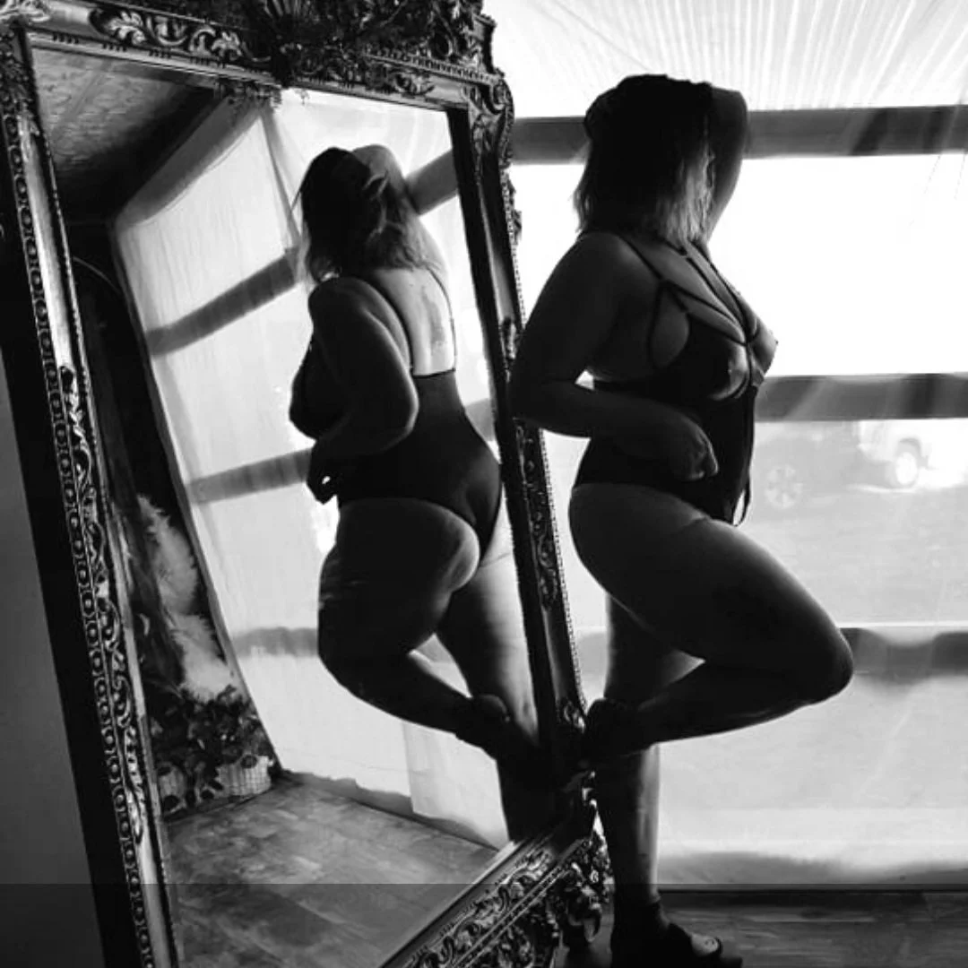 Person in black lingerie posing by an ornate mirror in black and white.