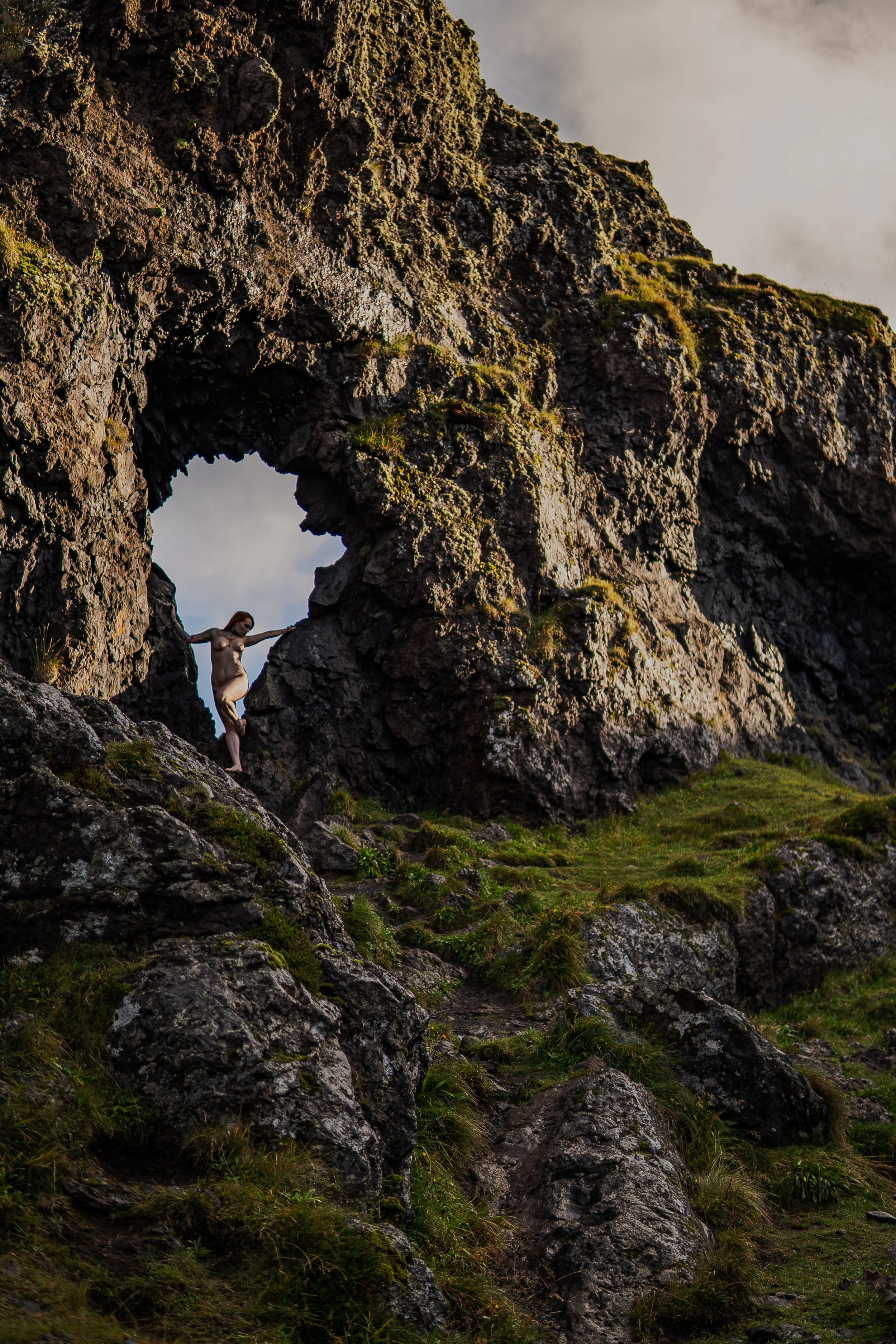 woman standing in a rocky arch formation on a grassy hillside in iceland during a boudoir photography session