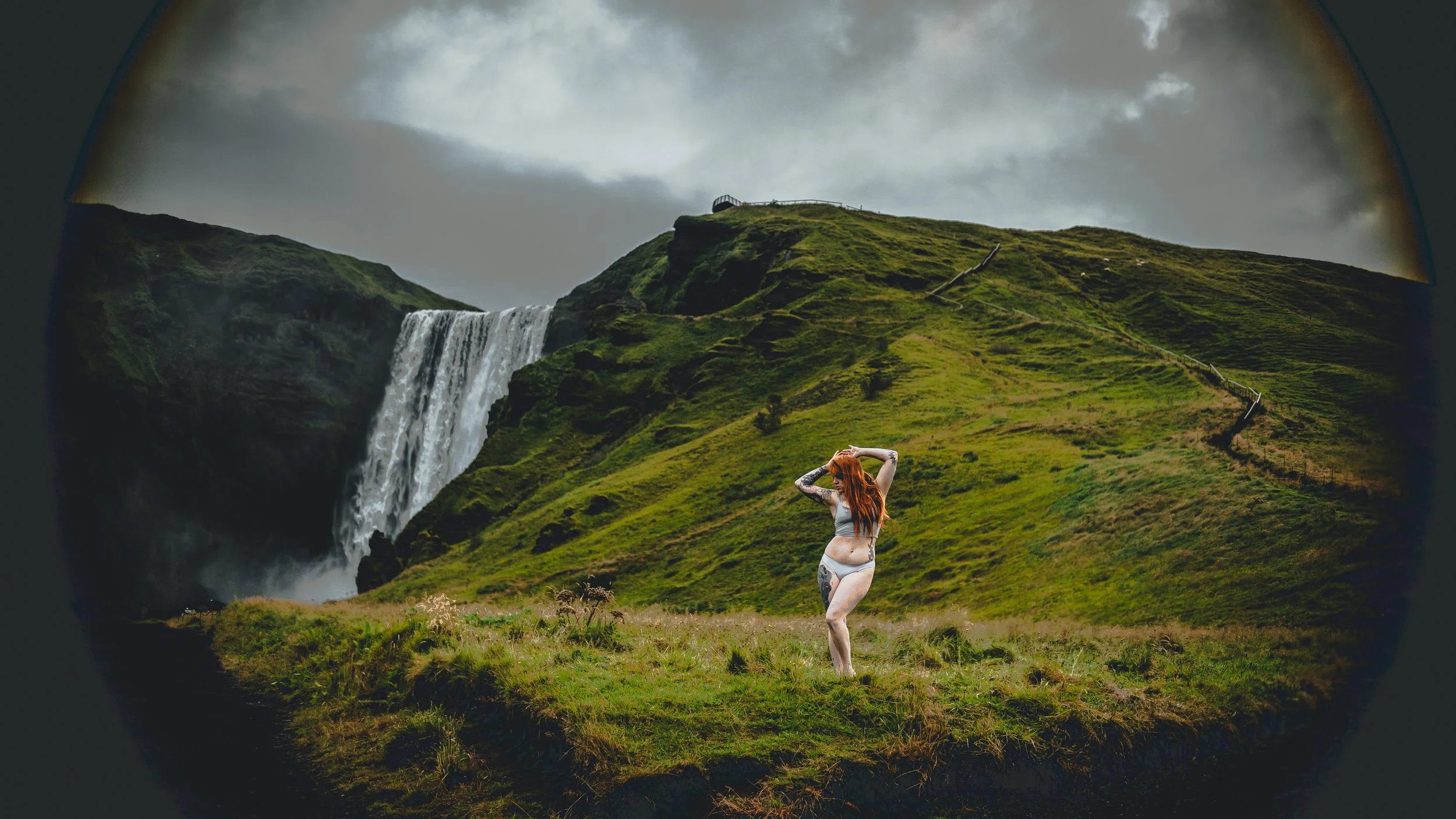 Woman standing in front of a waterfall and grassy hillside in iceland under a cloudy sky during a boudoir photography session