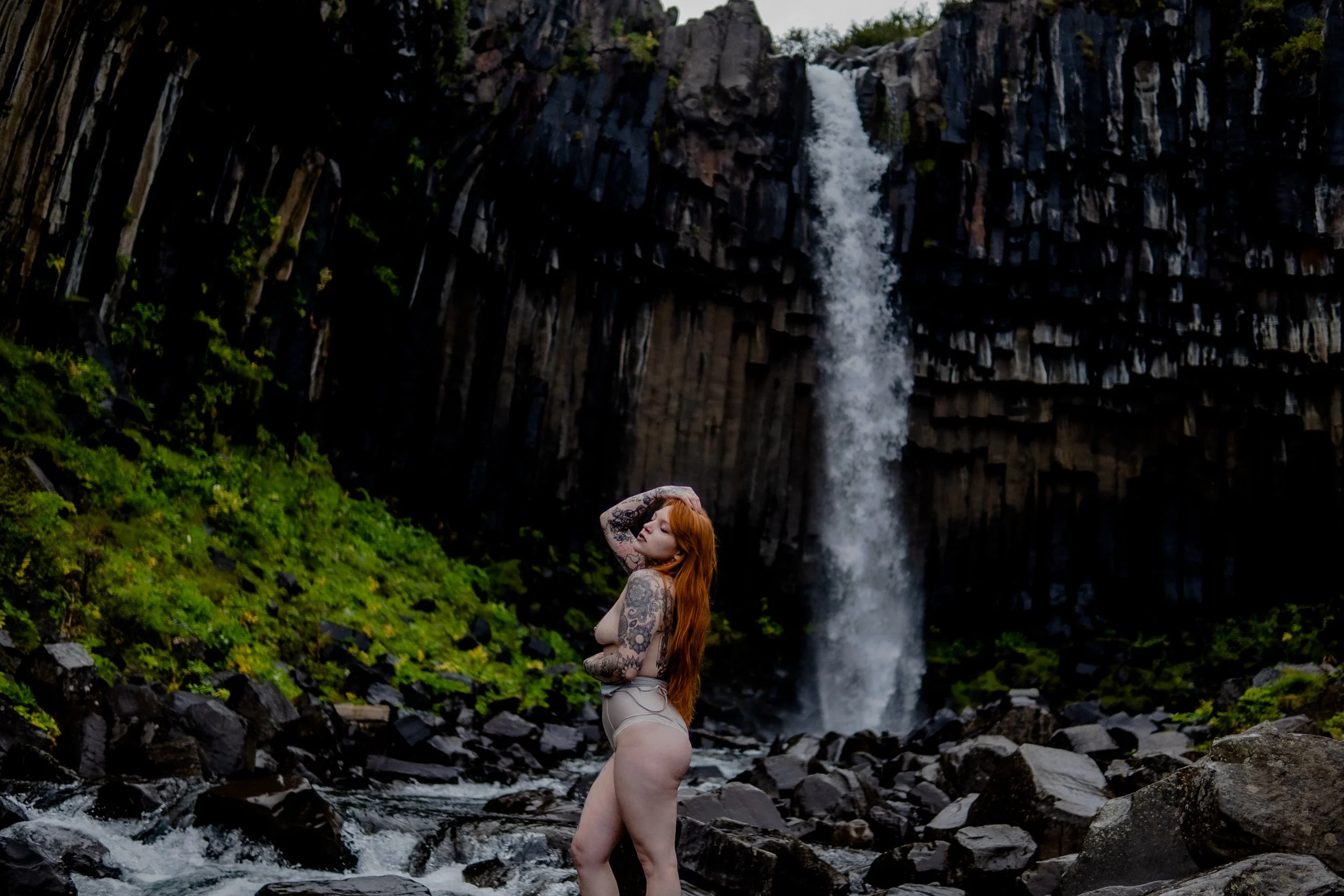 Person with tattoos and long hair standing in rocky landscape near waterfall in iceland during a boudoir photography session