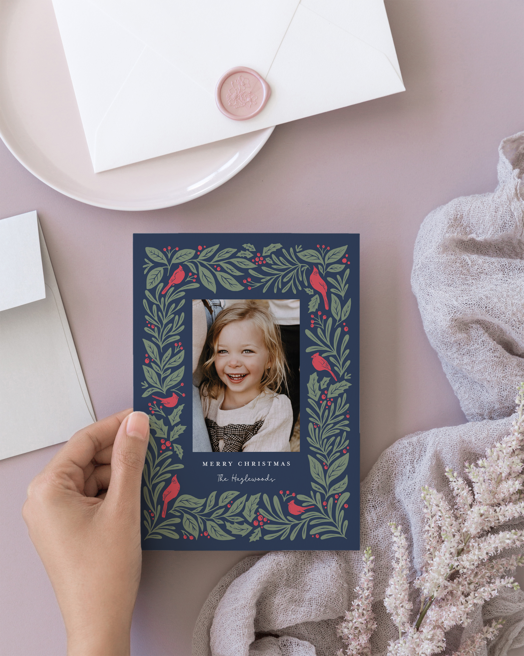 A person holding a Christmas card with a photo of a young girl smiling, surrounded by a decorative border of green leaves and red birds, with the text 'Merry Christmas' and 'The Hargrove' written below.