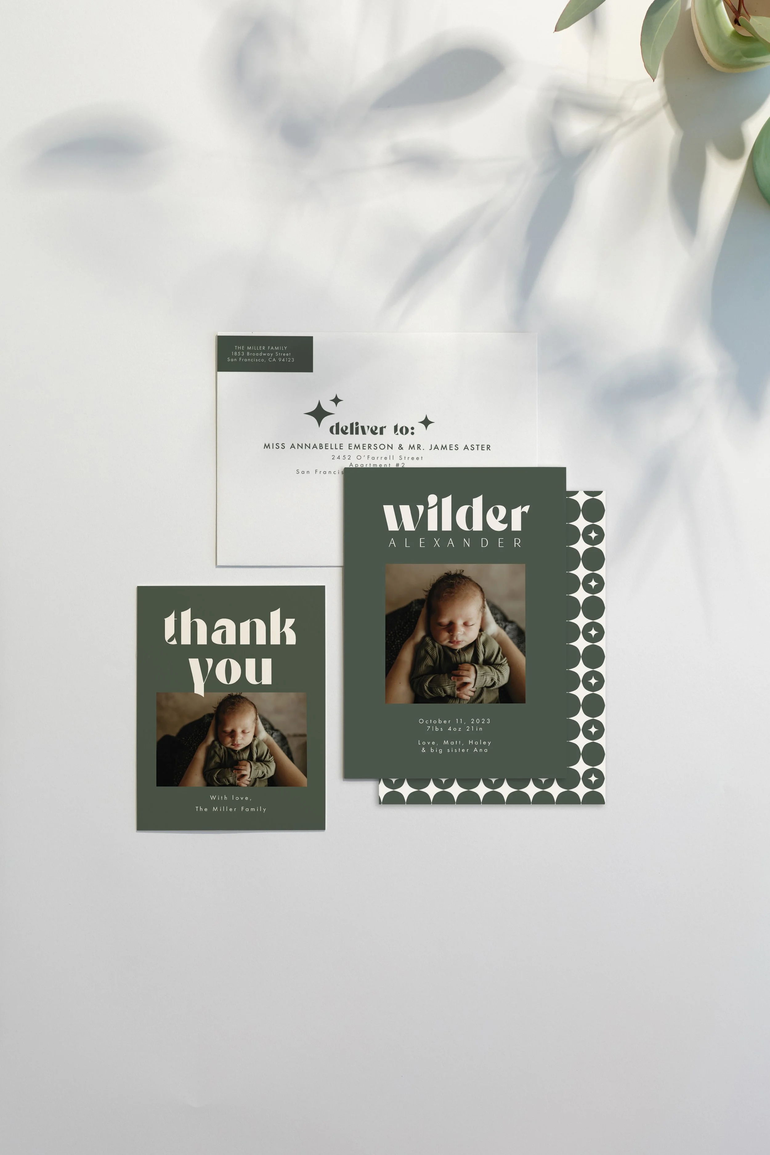 Baby announcement, thank you card, and envelope on a white surface with a plant in the background. The cards feature a photo of a sleeping baby and green and white text.