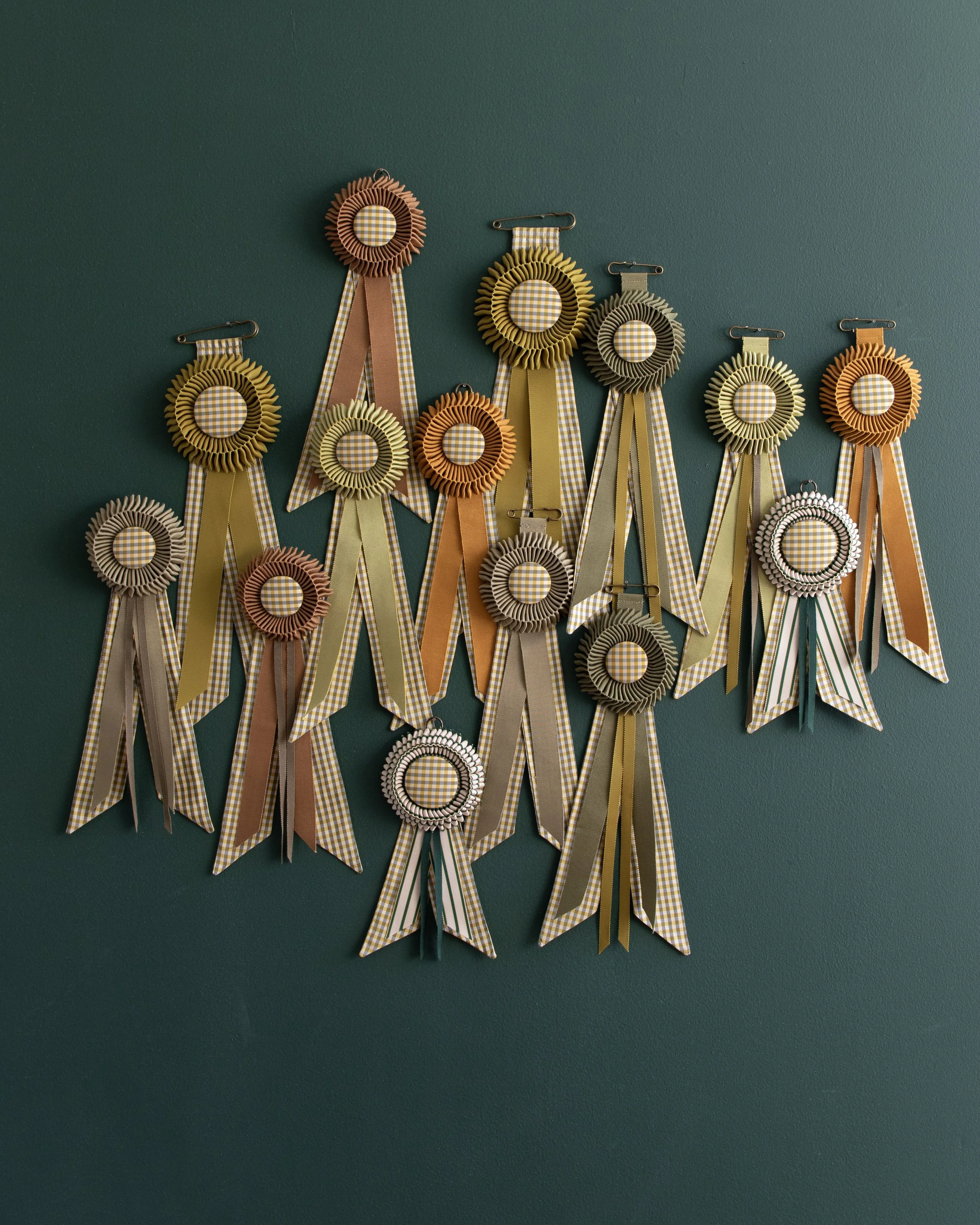 A collection of 14 rosettes made of petersham ribbon and silk taffeta on a dark green wall.