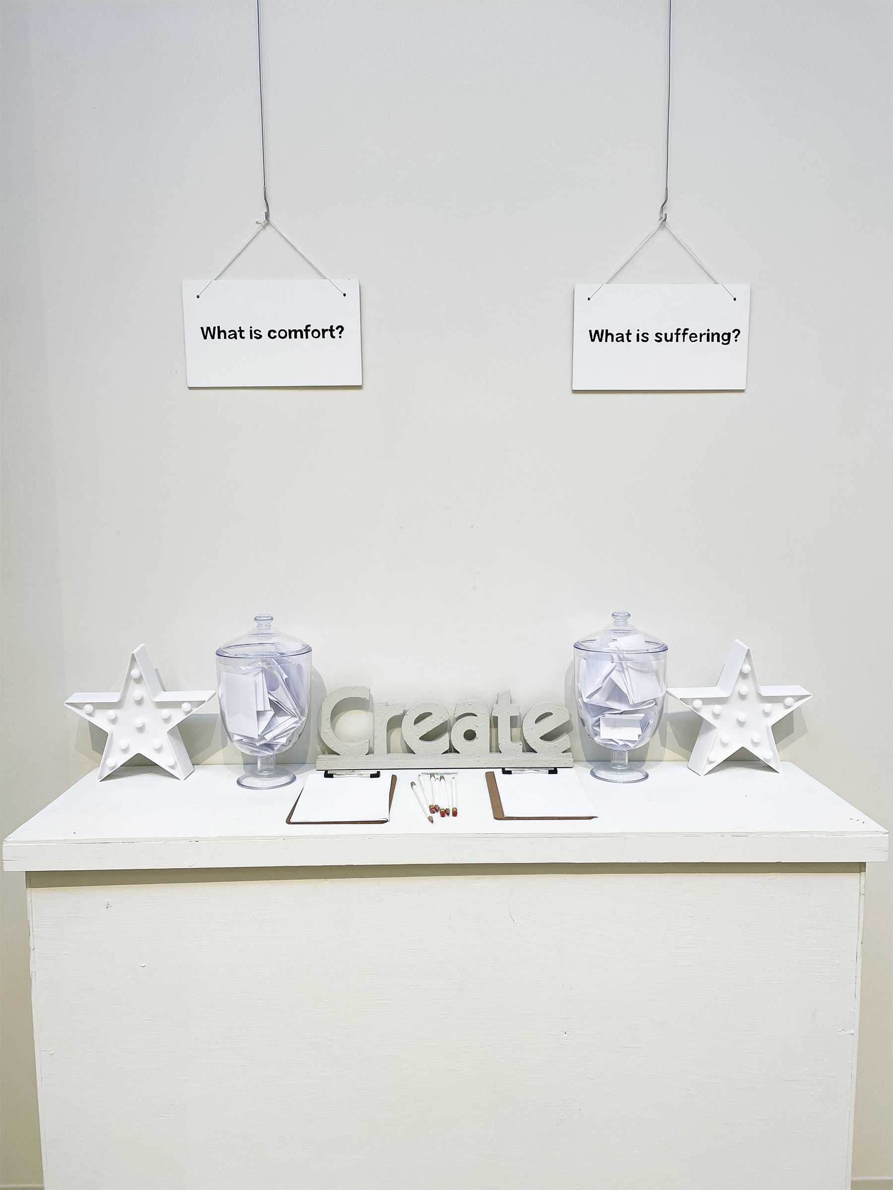 "What is Comfort? / What is Suffering?" 2016-present. Participatory Installation (wood signs, inspirational decor, plastic vessels, clipboards, pencils). Dimensions variable.