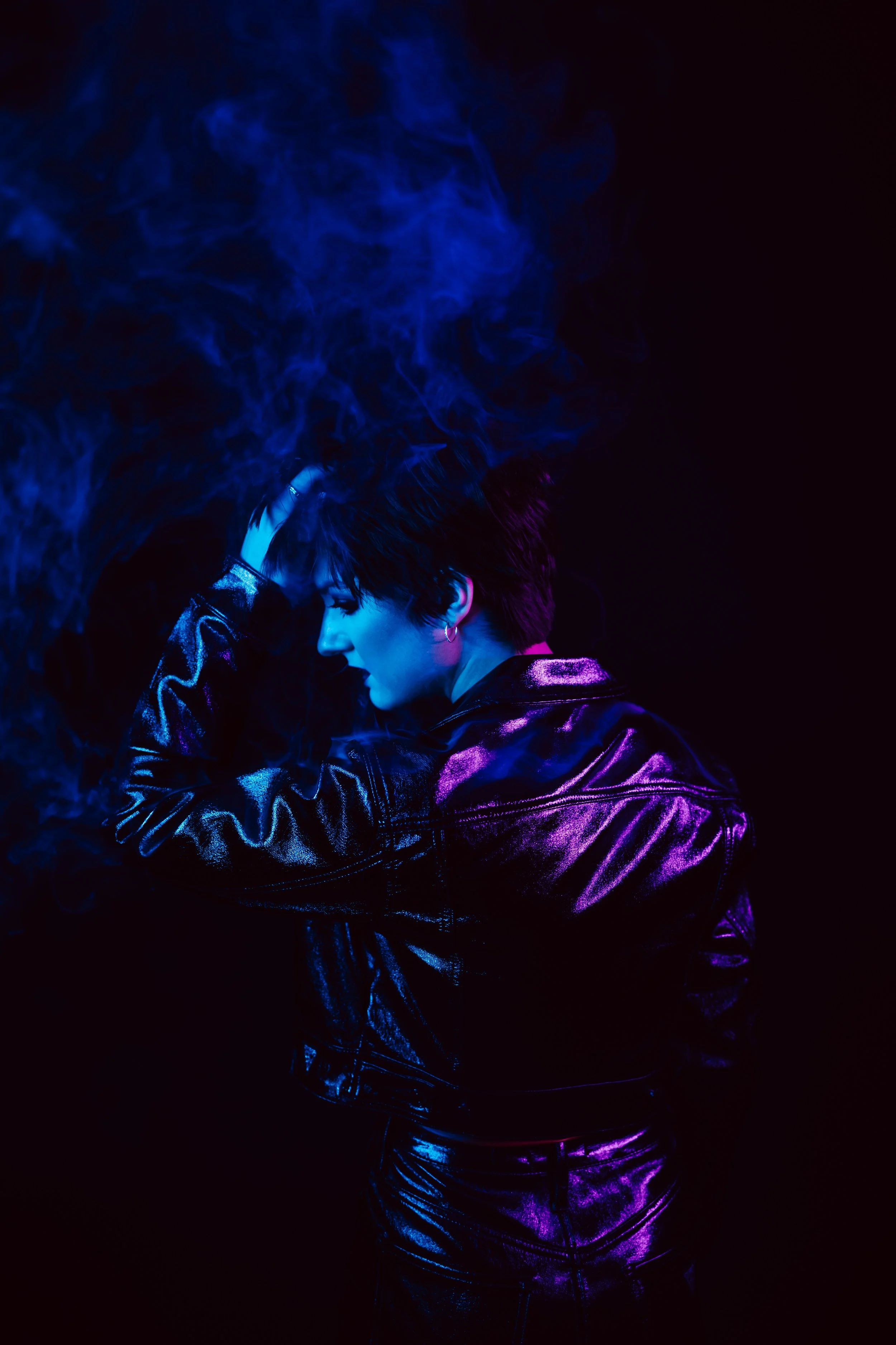 A woman with short dark hair and wearing a shiny black leather jacket is illuminated with blue and purple light, with blue smoke around her head, against a black background.