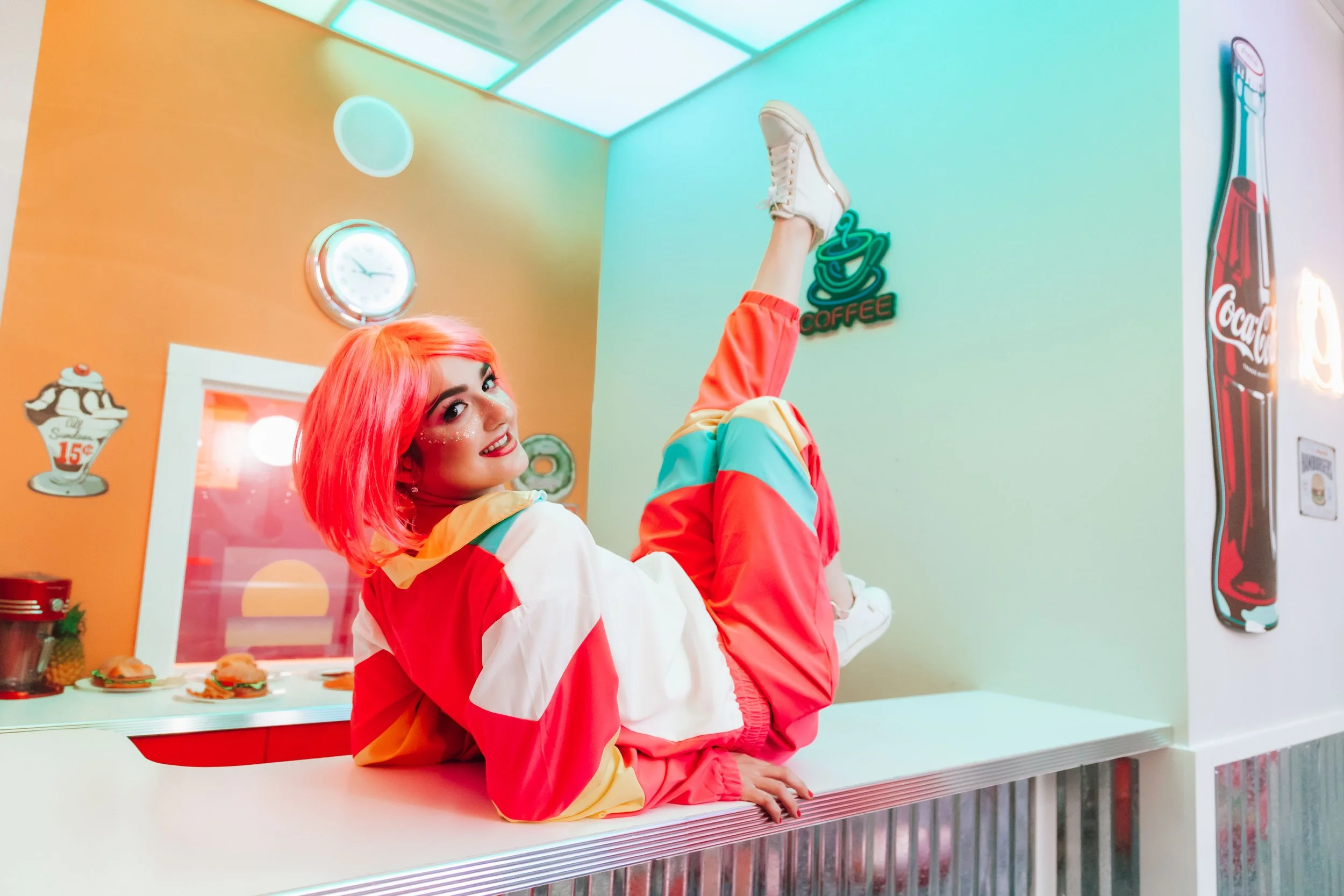 A woman with bright red hair lying on a diner counter, wearing colorful track pants and a jacket, with a retro-themed diner background including neon signs, clocks, and posters.