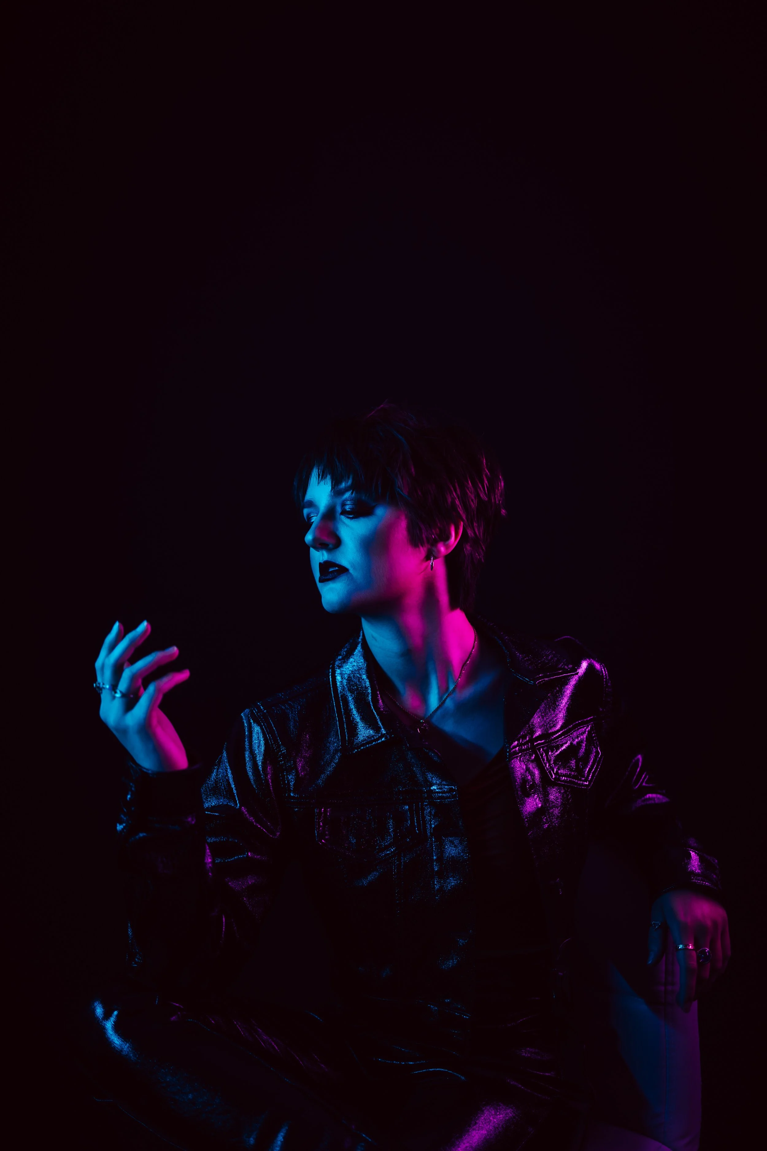 A woman with short hair and dark lipstick poses in a dark setting illuminated by multicolored lights, wearing a shiny black leather jacket and jewelry.