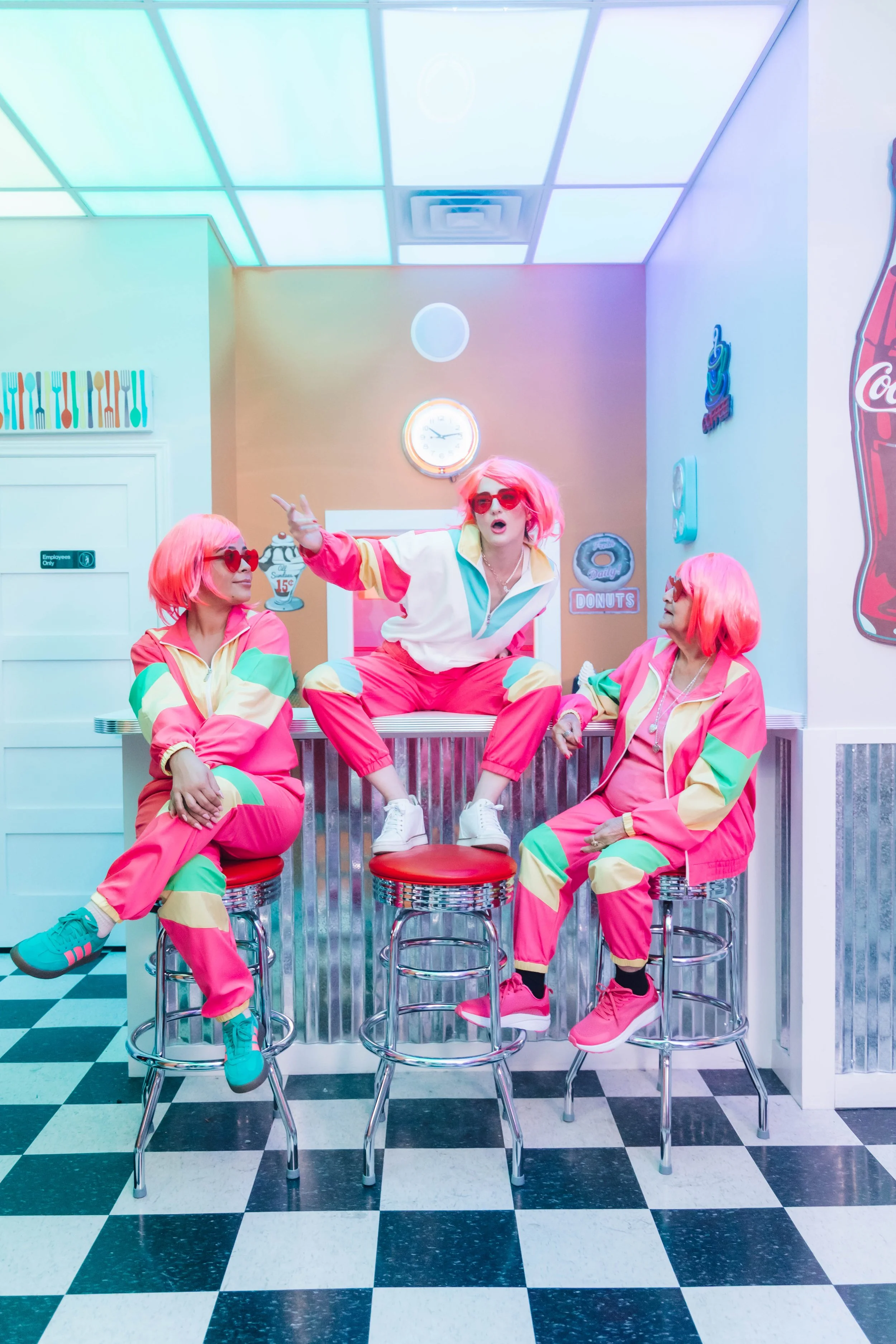 Three women with pink hair and retro outfits in a neon-colored diner, sitting on barstools and engaging in lively conversation.