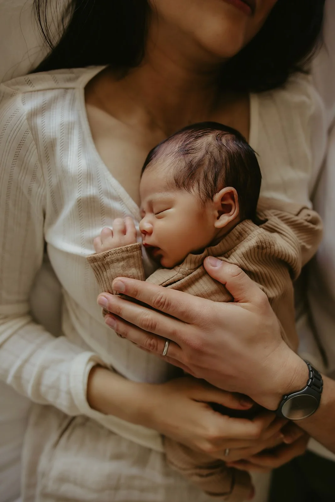A newborn baby sleeps on a woman's chest as an adult hand gently rests on the baby's back, beautifully captured by an in-home newborn photographer Sacramento.