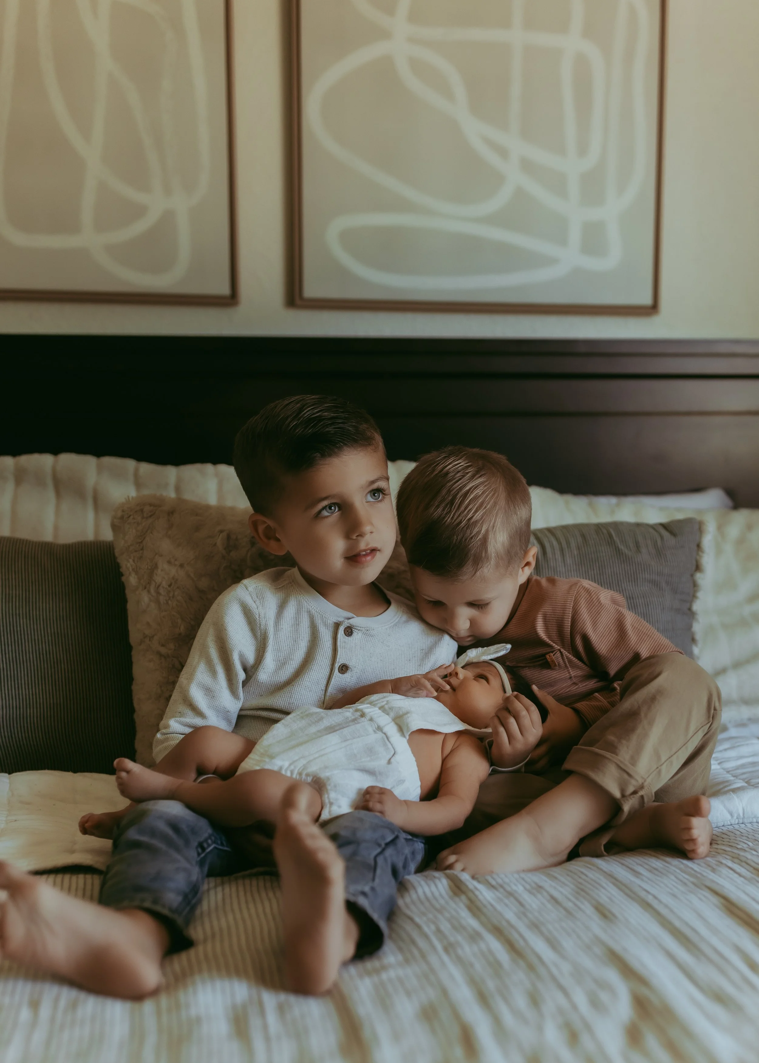  Two young boys sit on a bed, gently cradling a sleeping baby, with abstract art adorning the wall behind them. Captured by a Roseville Newborn photographer, this tender moment encapsulates the serene beauty of new beginnings. 