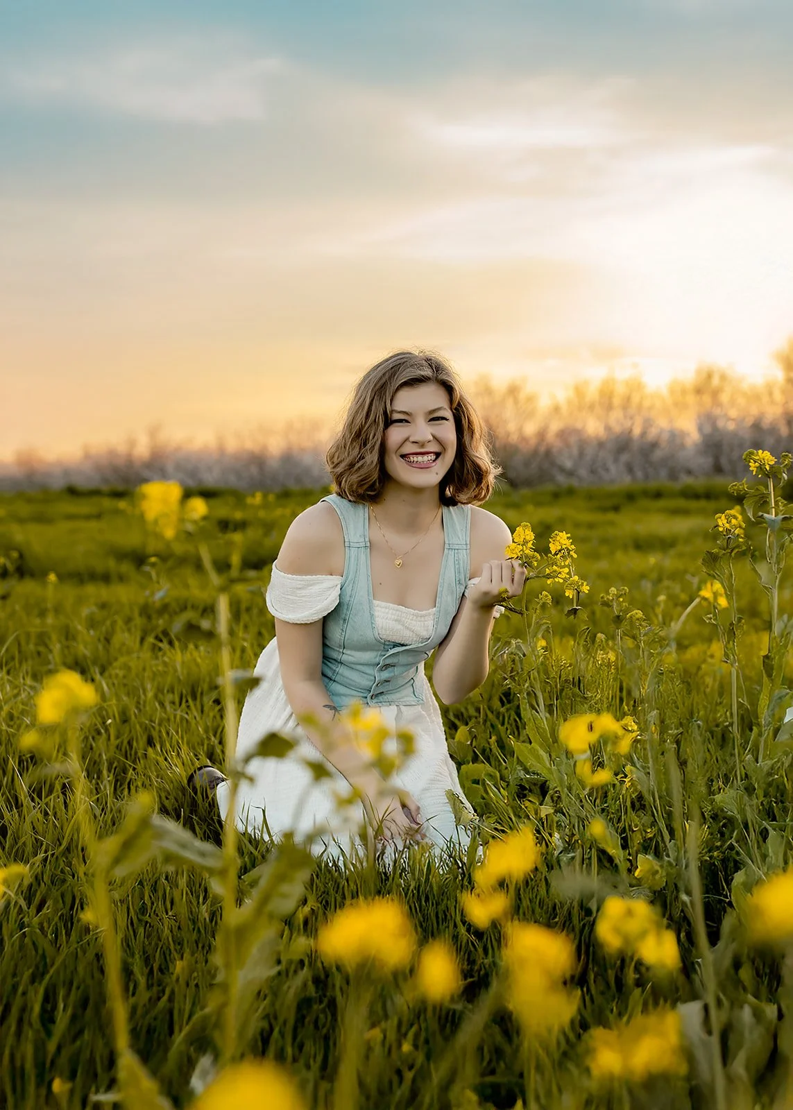 Woman in a white dress kneels in a field of yellow flowers at sunset, smiling at the camera—captured by a Sacramento senior photographer specializing in high school senior photography Sacramento.