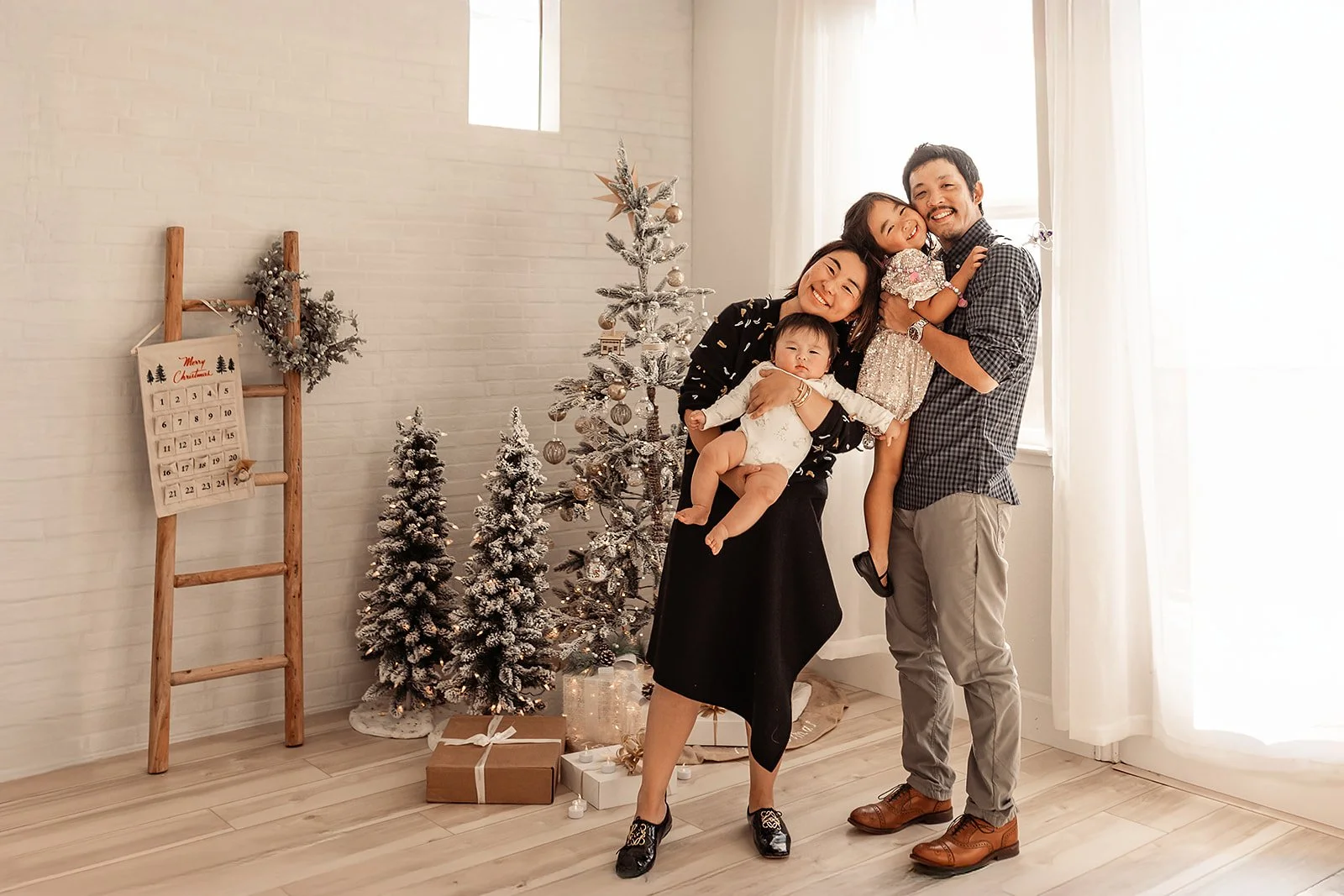 A family of four smiles joyfully, posing in a festive room decorated with Christmas trees and a wooden ladder, beautifully captured by a talented Roseville photographer.