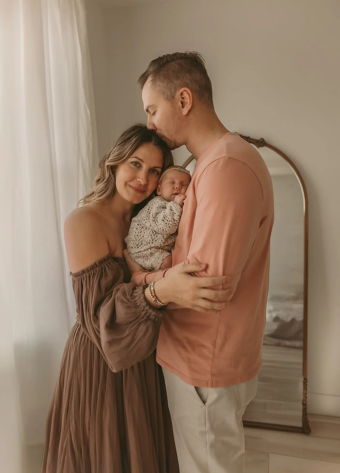 A couple lovingly embraces their newborn baby in a softly lit, cozy room with a mirror in the background—perfectly capturing the warmth of an in-home newborn photographer Sacramento session.