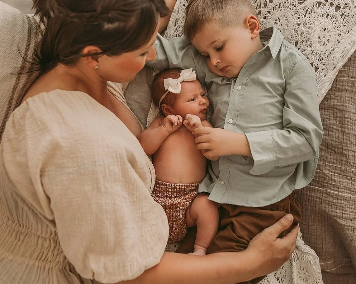  A woman cradles a baby and young child, nestled closely with a lace backdrop, beautifully captured by a Roseville Newborn photographer. 