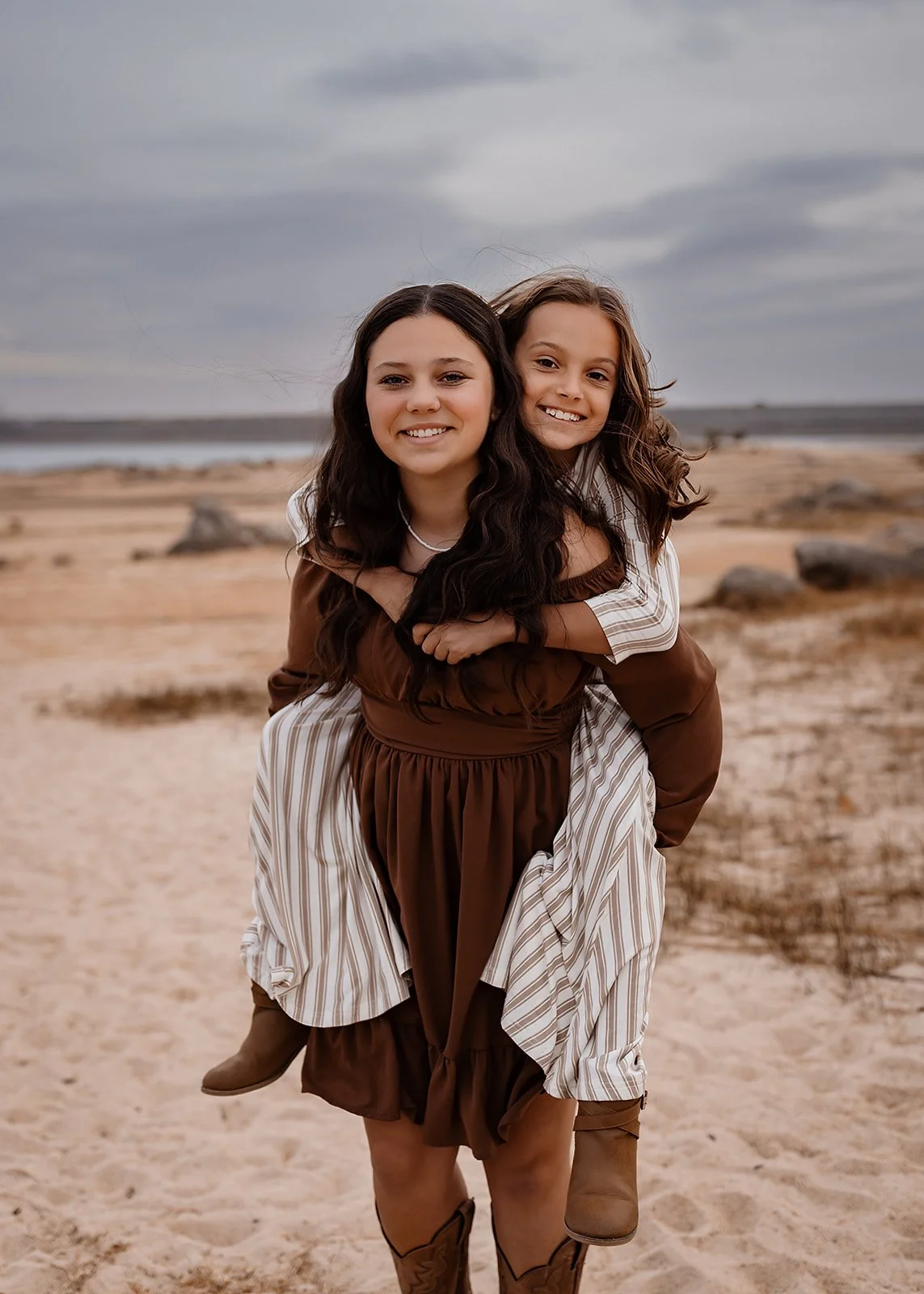  A girl gives another girl a piggyback ride on a sandy beach, both smiling under a cloudy sky, capturing the essence of Northern California family photography. 