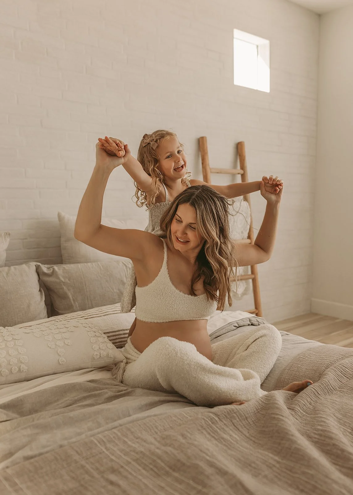 A woman in matching loungewear sits on a bed in El Dorado Hills, a smiling child joyfully perched on her shoulders, holding her hands—a perfect moment captured by an El Dorado Hills maternity and motherhood photographer.