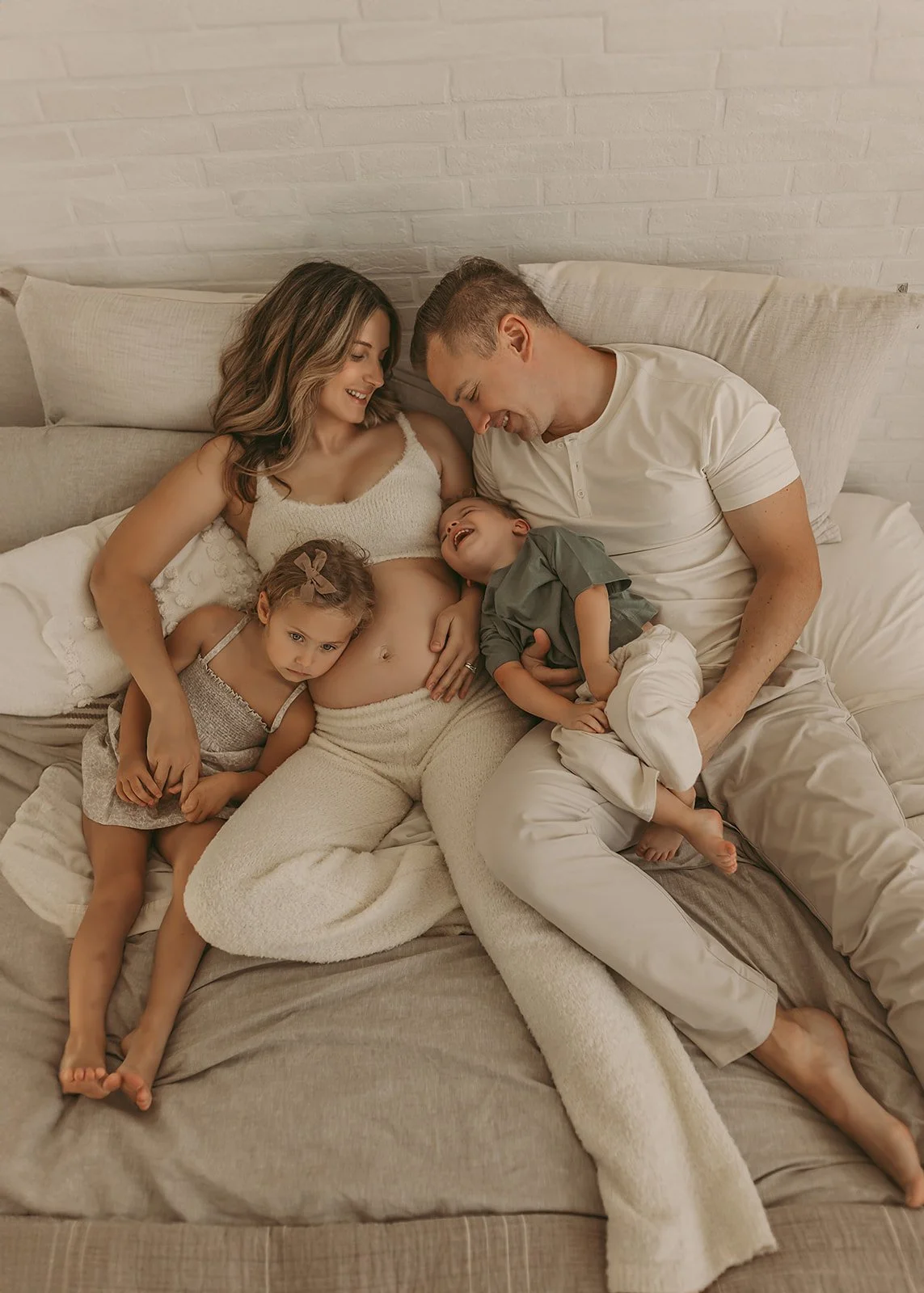 A pregnant woman lies on a bed, smiling with her partner and two young children cuddling beside her—captured by an in-home newborn photographer Sacramento.