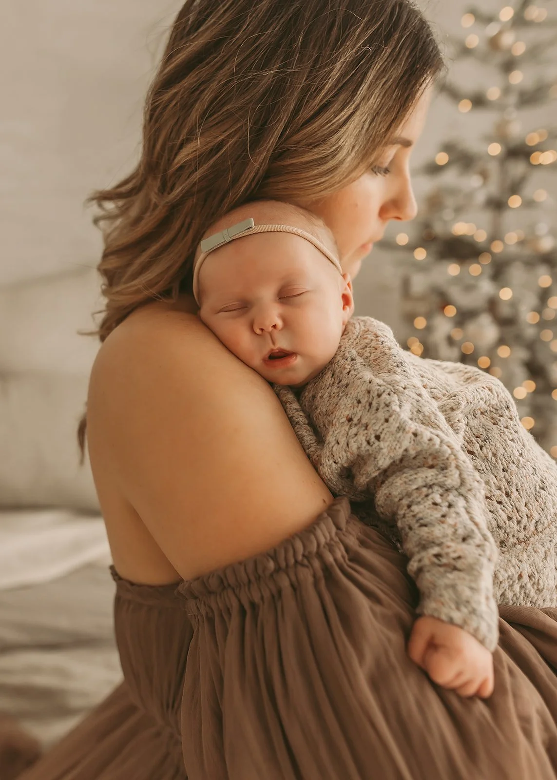 A mother holds a sleeping baby on her shoulder, with a lit Christmas tree in the background—captured by an in-home newborn photographer in Sacramento.