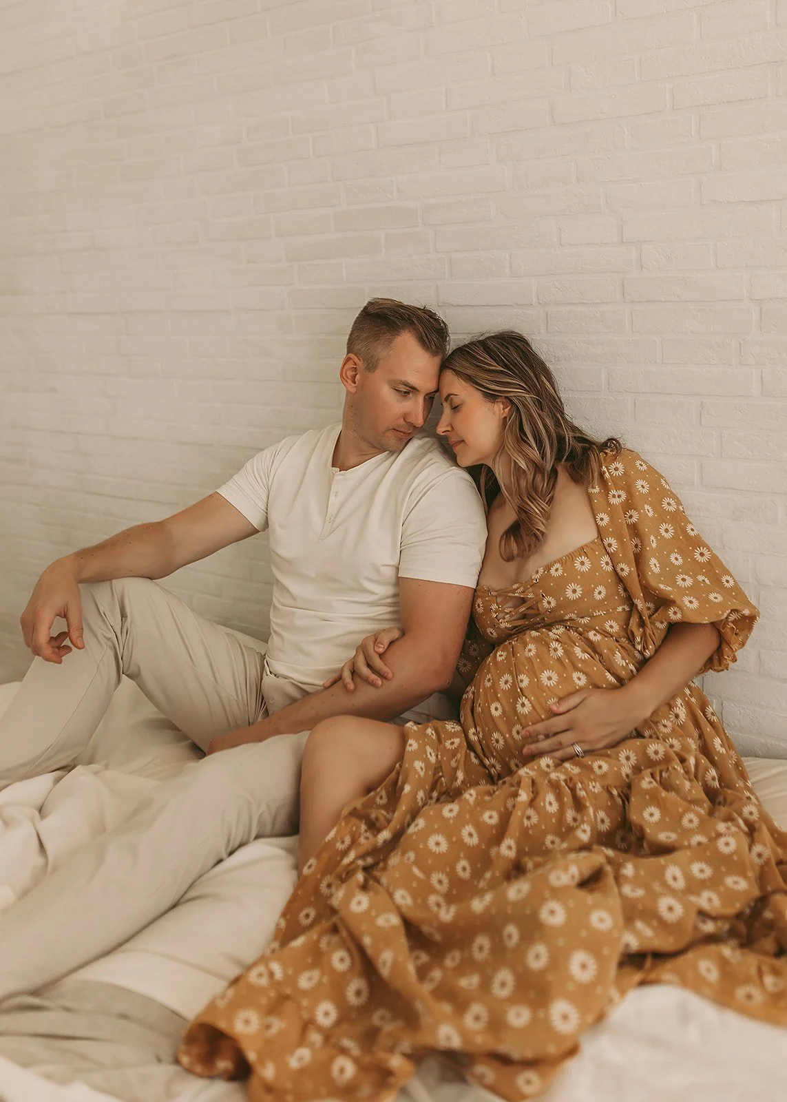 A couple sits closely on a bed; the woman is pregnant and wearing a floral dress, both look peaceful—a perfect moment captured by a Sacramento maternity and newborn photographer.