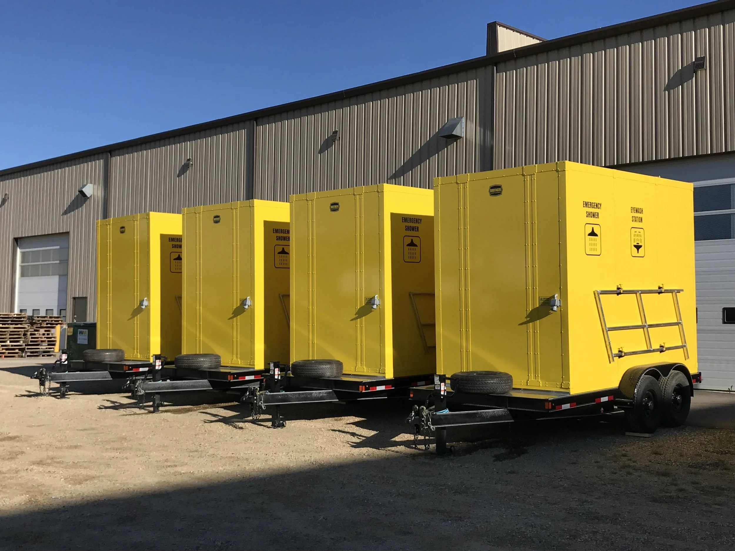 Four yellow emergency safety shower and eyewash trailer stations parked outside a warehouse with shower/eyewash icons.