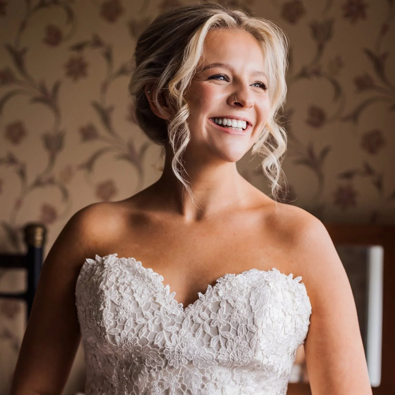 A smiling woman with blonde hair styled in loose curls, wearing a strapless white lace wedding dress, standing indoors with floral wallpaper in the background.