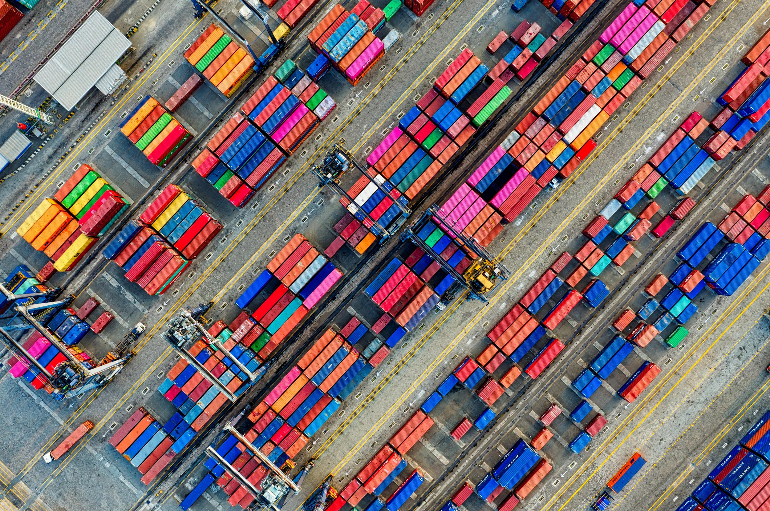 An aerial view of a large shipping yard filled with colorful cargo containers arranged in rows, with cranes and trucks moving containers.