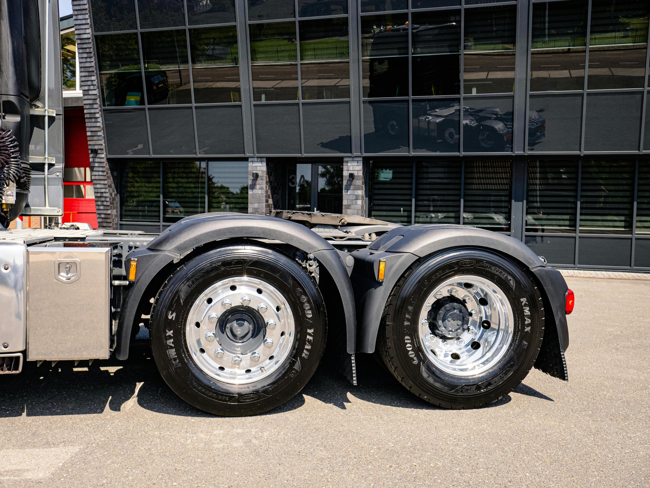 Close-up of the rear wheels and tires of a semi-truck parked outside a modern building with glass windows.