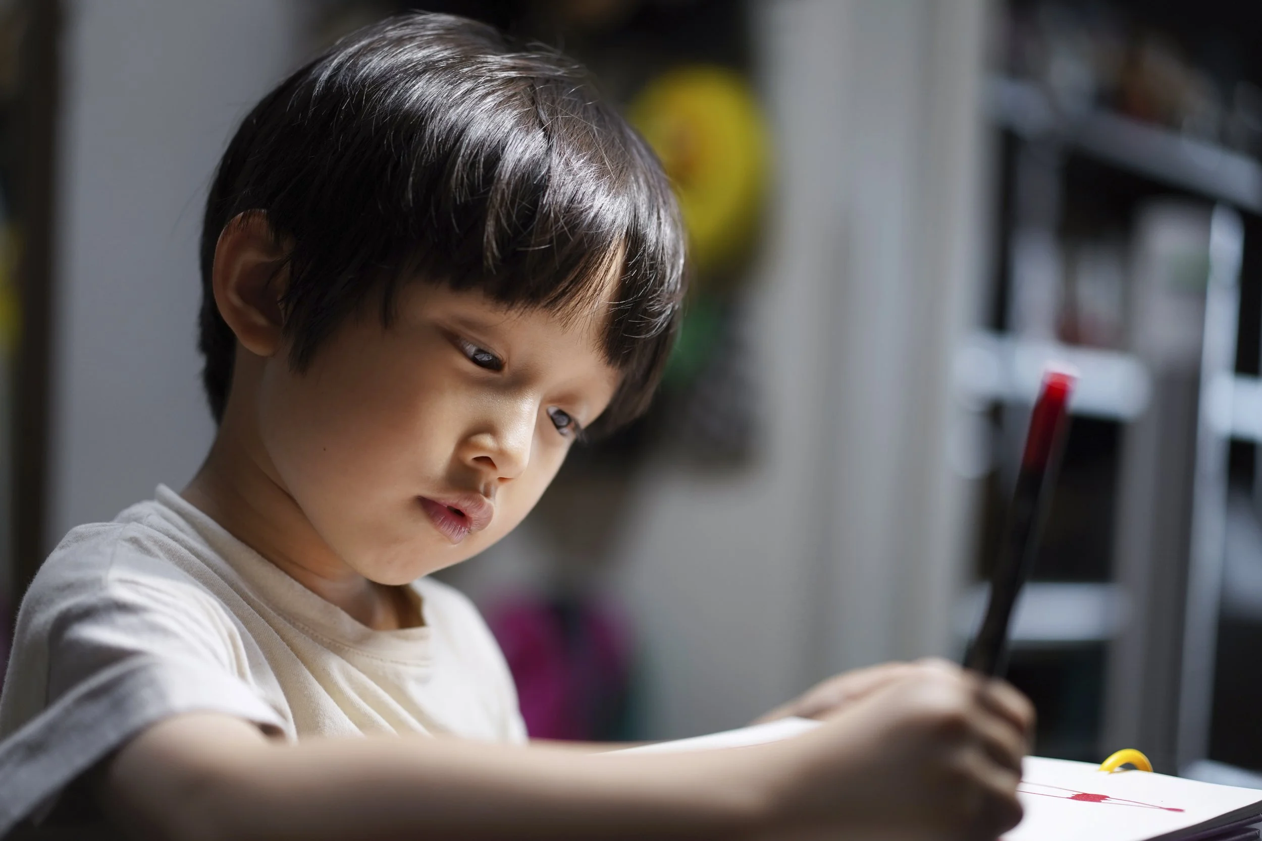 A young child with dark hair drawing or writing with a red marker on paper, indoors with shelves and a yellow balloon in the background.
