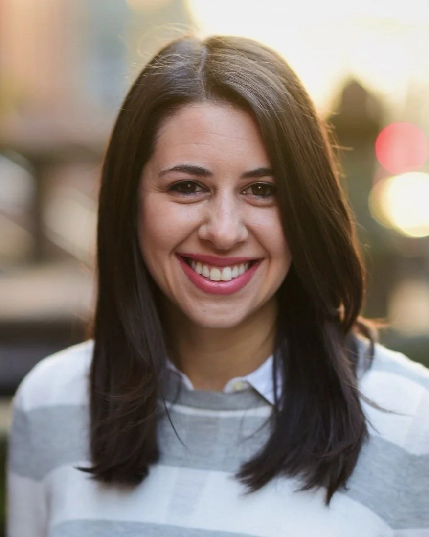 A young woman with dark brown hair and light skin smiling outdoors during sunset.