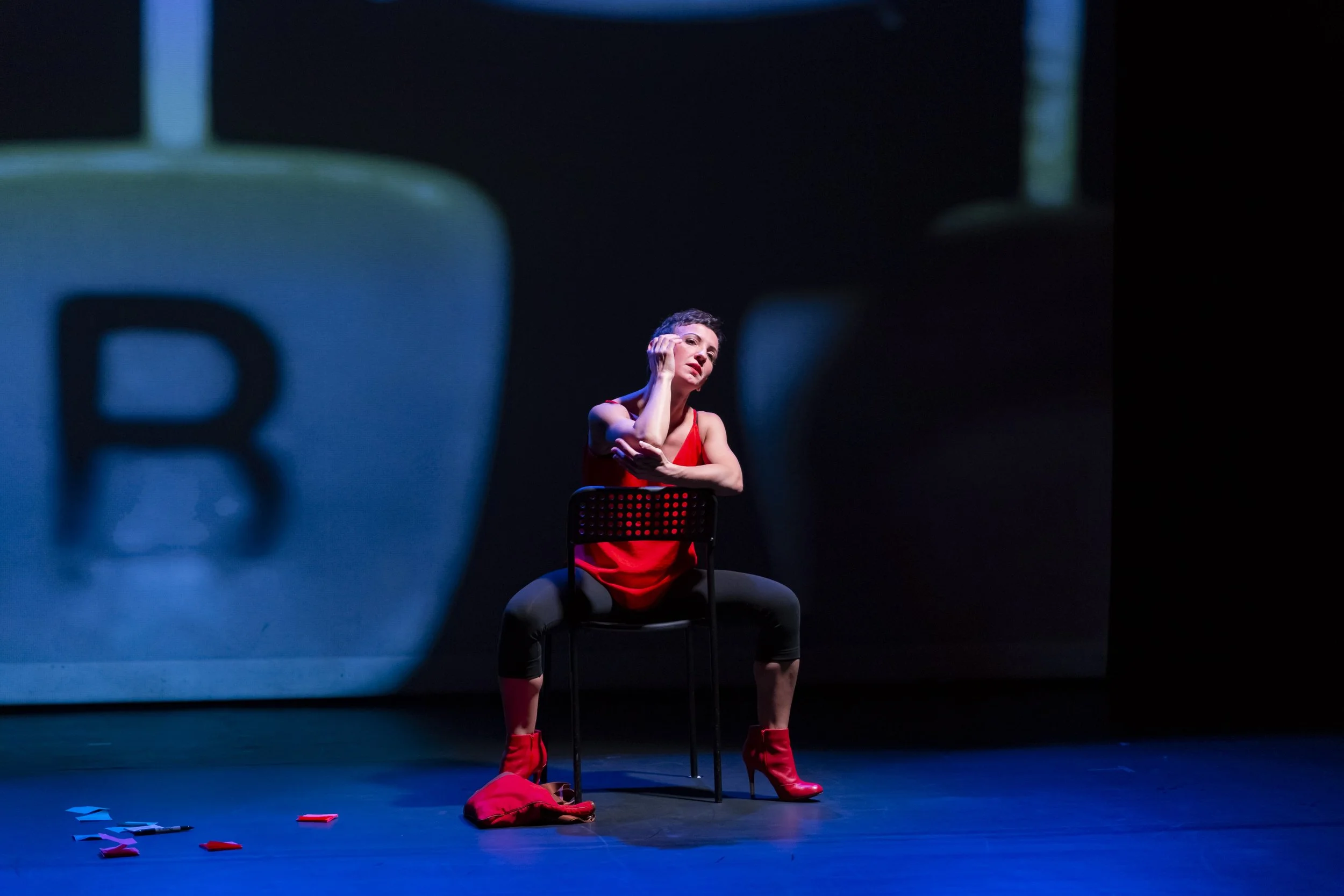 Valentina Priolo, performing artist sits on a chair on stage during a dance theater performance.