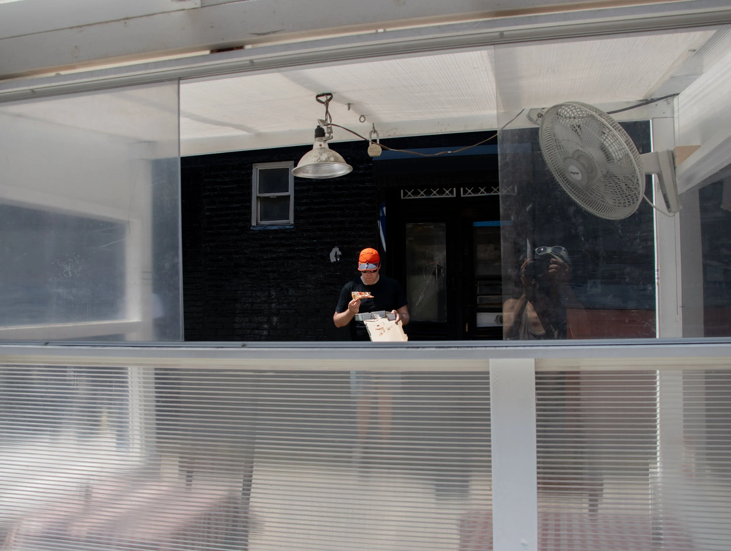 Person in black shirt, orange packable Parapack P-CAP, and sunglasses looking at pizza box while eating pizza, viewed through a reflective surface, with a wall fan and light fixture above, and photographer holding camera visible in reflection.