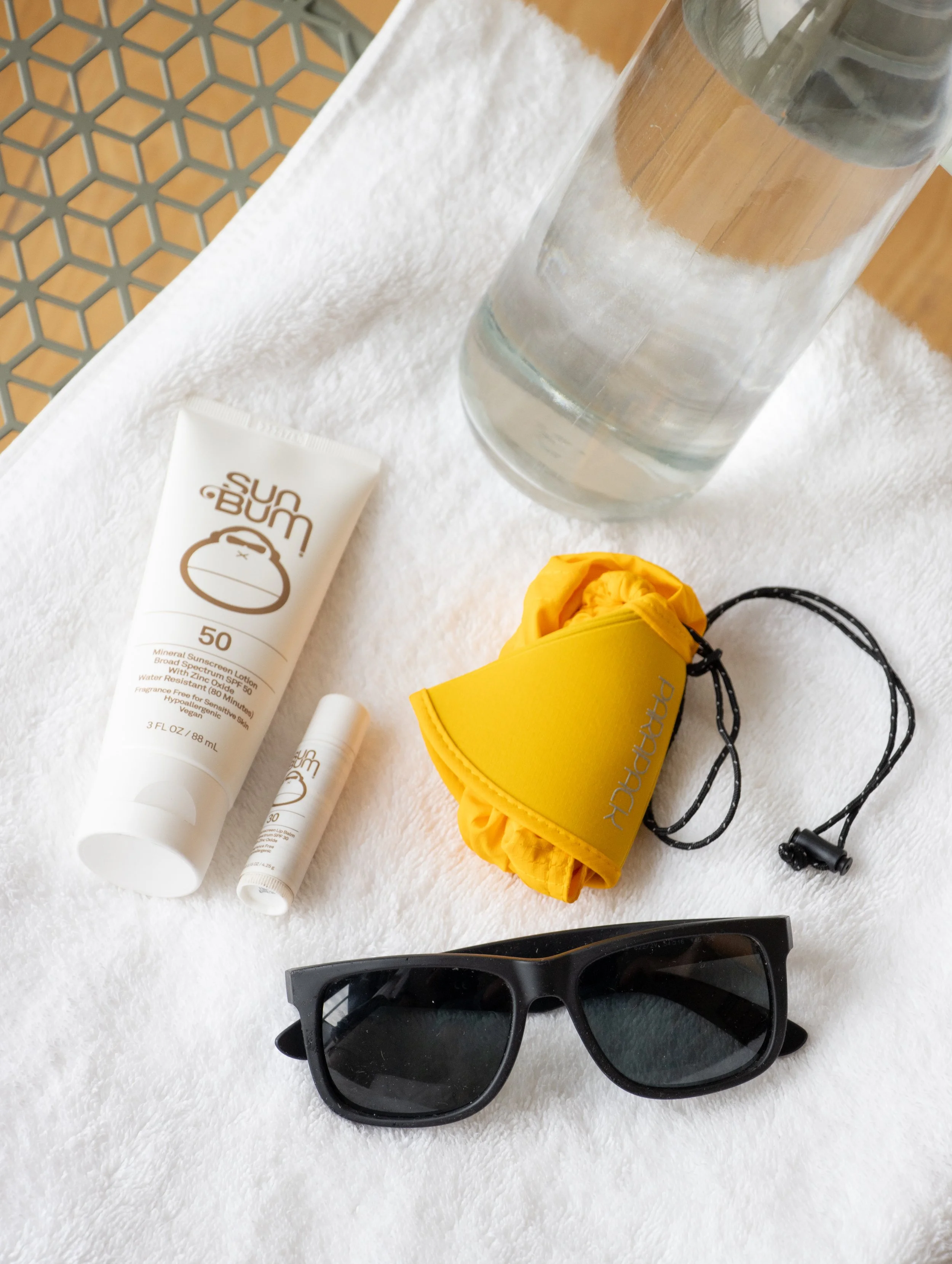 Sunscreen, lip balm, yellow Parapack P-CAP in compact mode, black sunglasses, and a water bottle on a white towel.