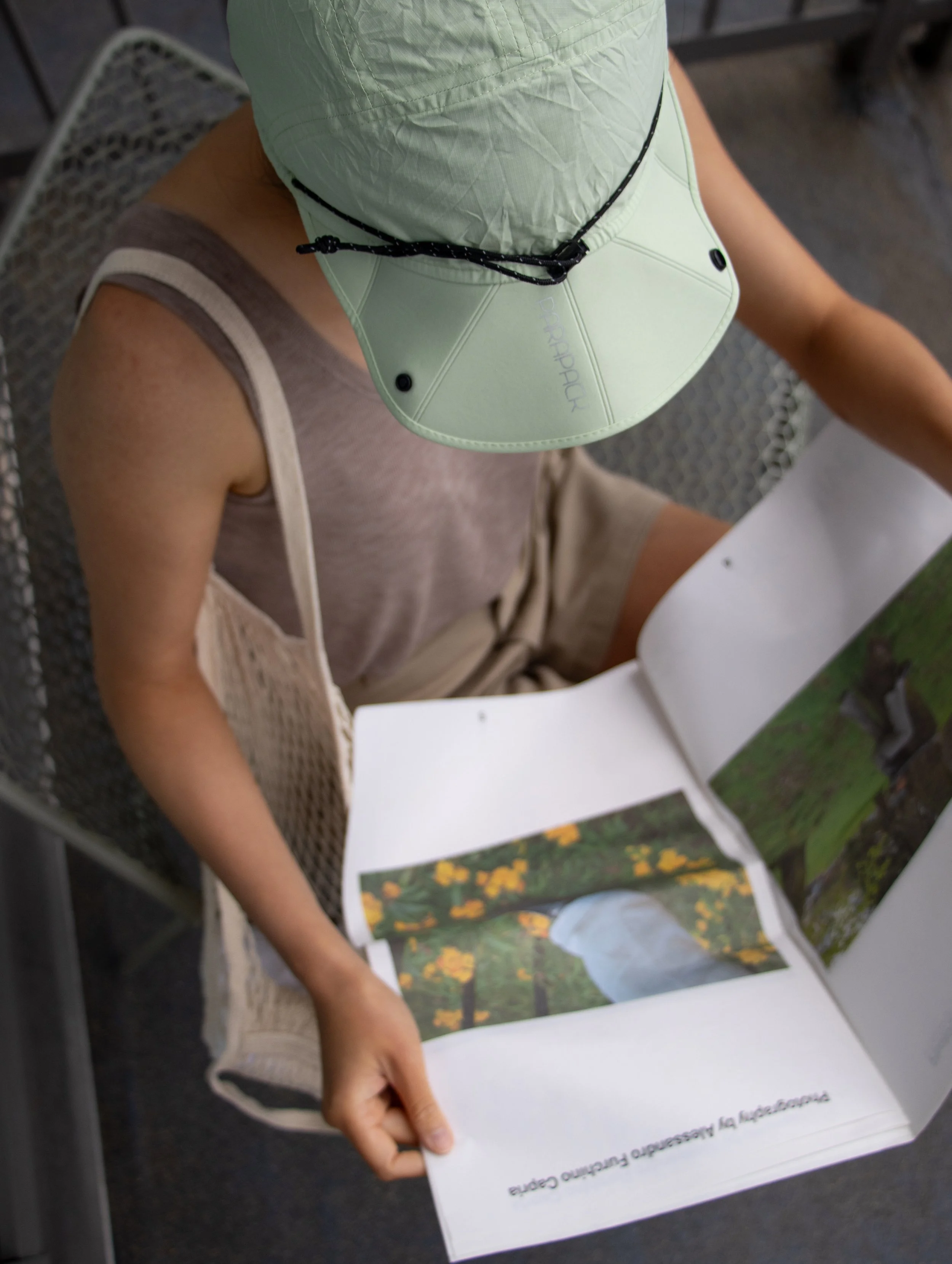 A person wearing a light green packable Parapack P-CAP sitting at a metal table, looking at a photo book with pictures of flowers and greenery.