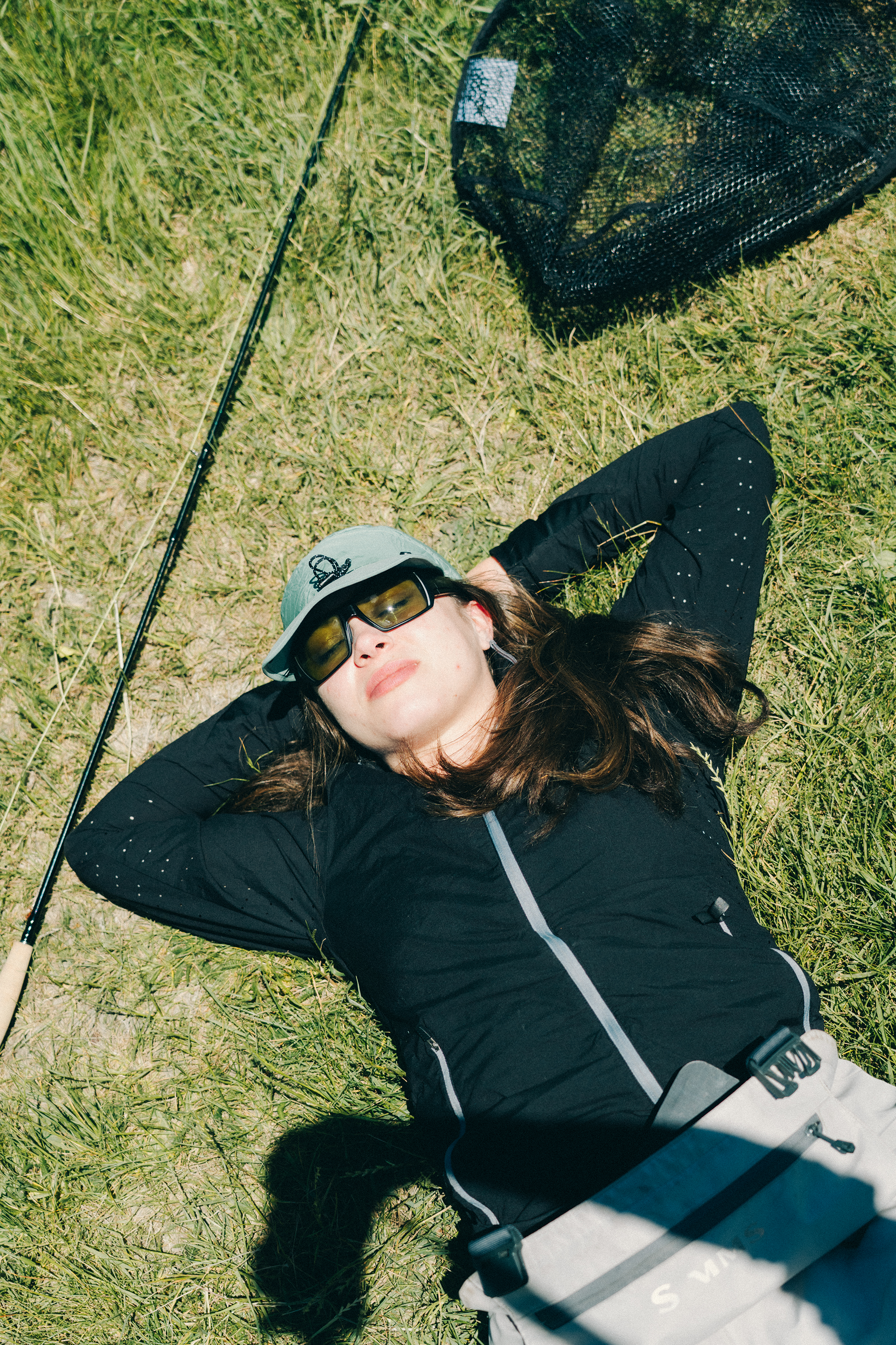 A person laying down on the grass taking a nap under the sun, her fishing pole and net are besides her. 