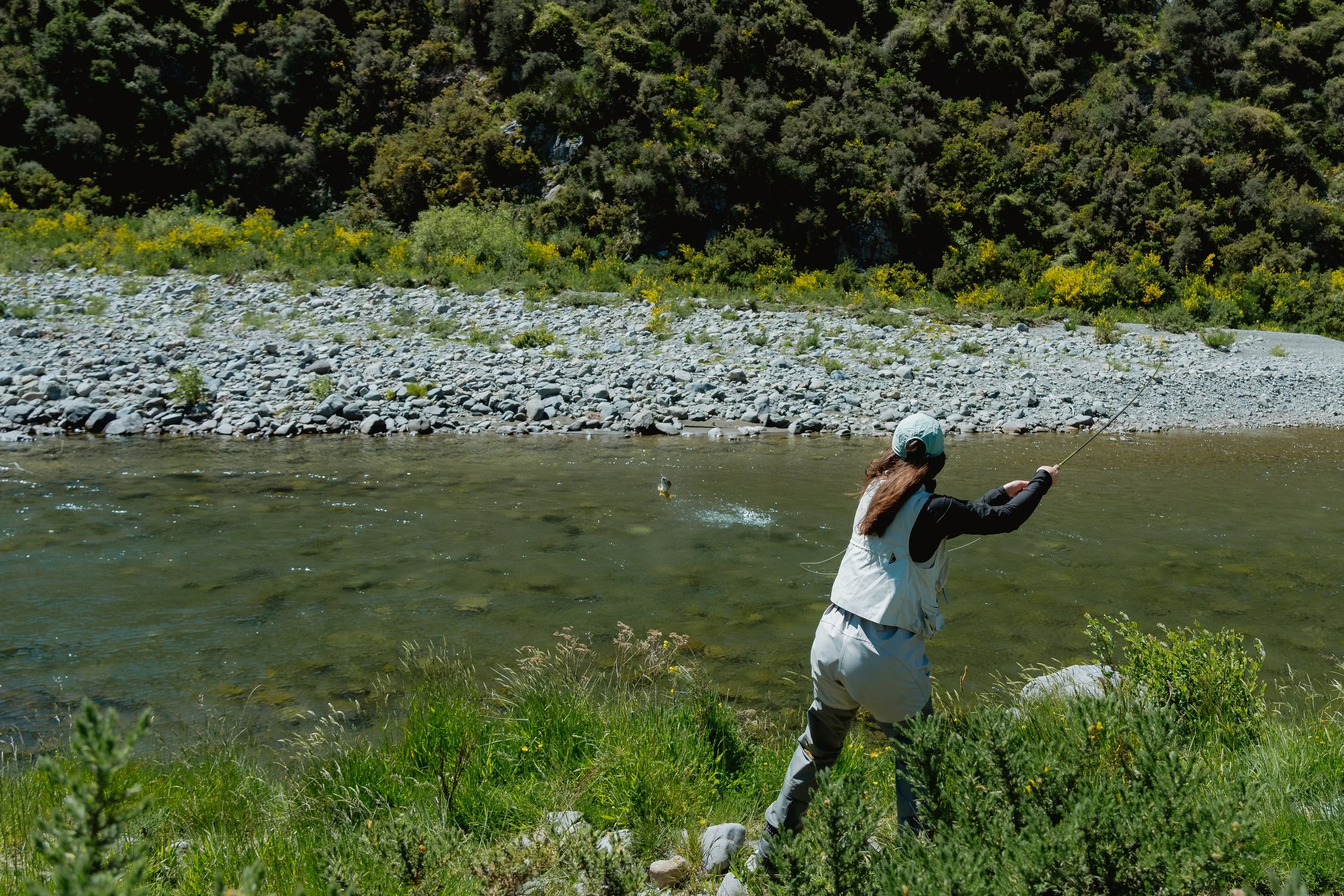 A person fishing in a river surrounded by greenery, rocks, and hills under sunny weather.