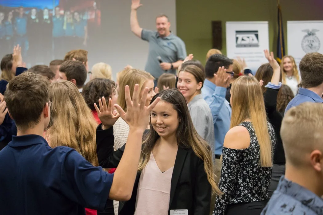 A diverse group of young people participating in a classroom activity, raising their hands, with an instructor smiling and waving at the back, in a professional setting with banners and a projected slide in the background.