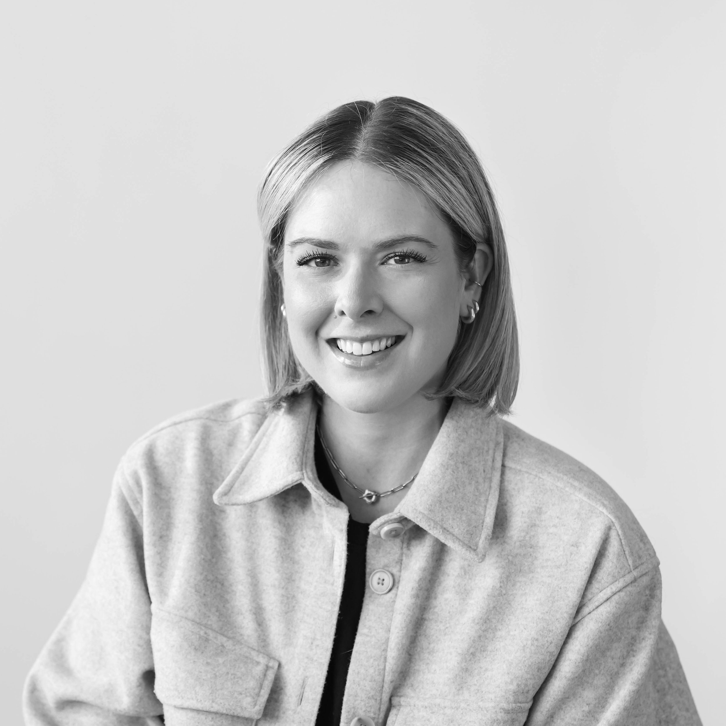 Black and white photo of a woman's face, smiling with her teeth showing, wearing a light-colored coat, earrings, and a chain necklace, against a plain background.