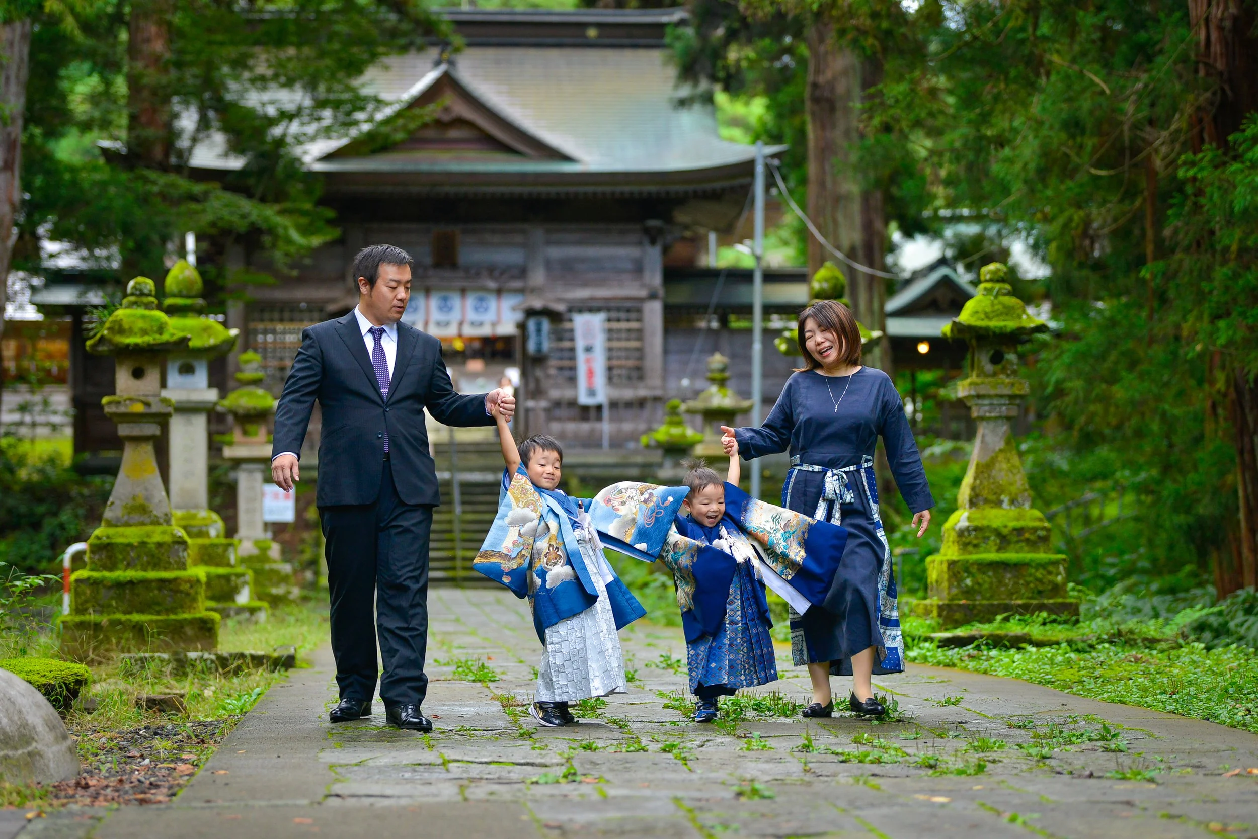 家族が神社の境内を歩きながら子供たちを祝福している写真。子供たちは着物を着ており、家族はカジュアルな服装。背景には古い木造の神社と苔むした灯篭が見える。