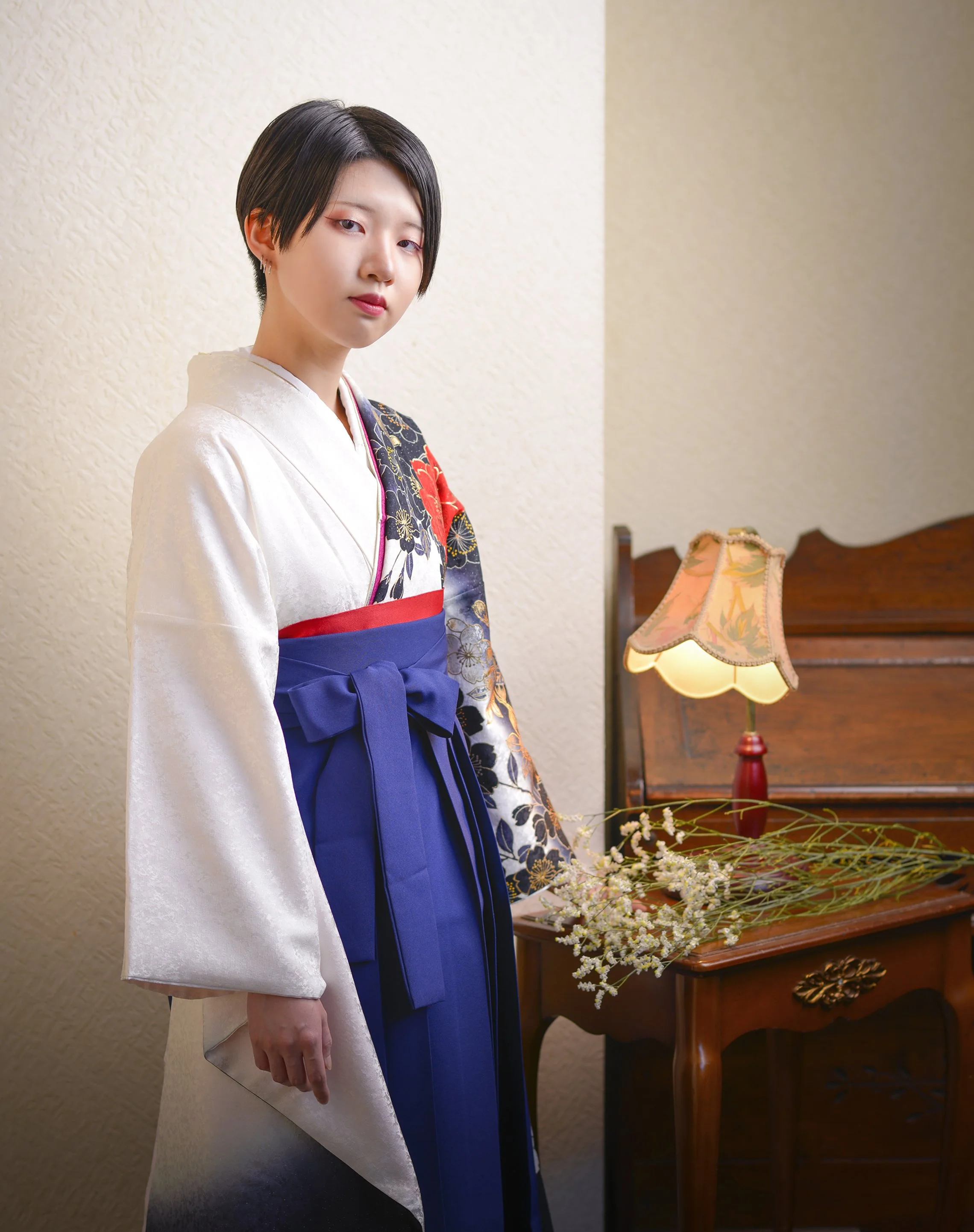 伝統的な着物を着た若い女性が室内に立っている。背景には木製の家具とテーブルの上に花とランプが置かれている。