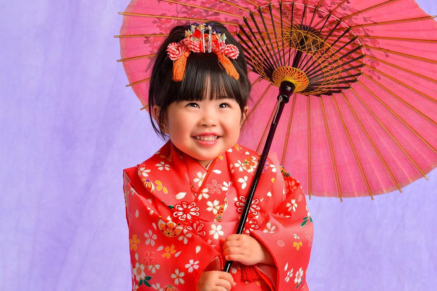 ピンク色の和服を着た若い女の子が、七五三の着物を着て、和傘を持ち、微笑んでいる写真。