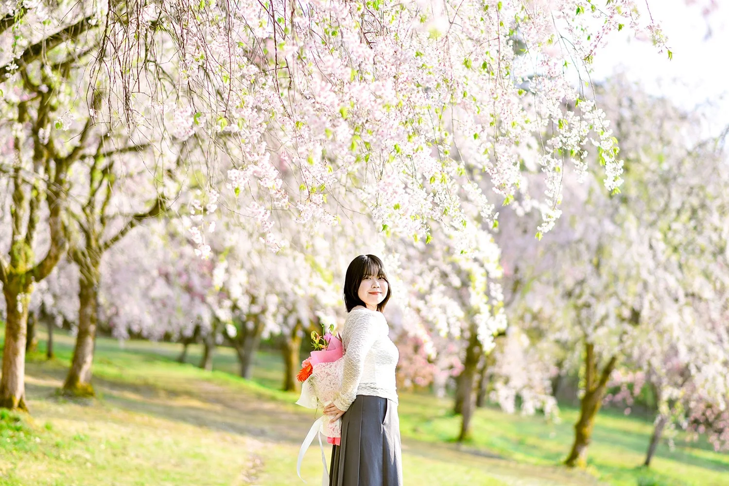 桜の花が咲く公園を背景に、花束を持った女性が微笑んでいる風景.