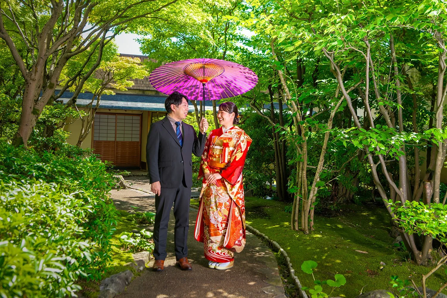 人々が伝統的な着物とスーツを着て、お店の前の庭園を散策している様子。女性は赤い着物を着て、男性は黒いスーツを着ている。女性は紫色のパラソルを持ち、二人は笑顔で話している。